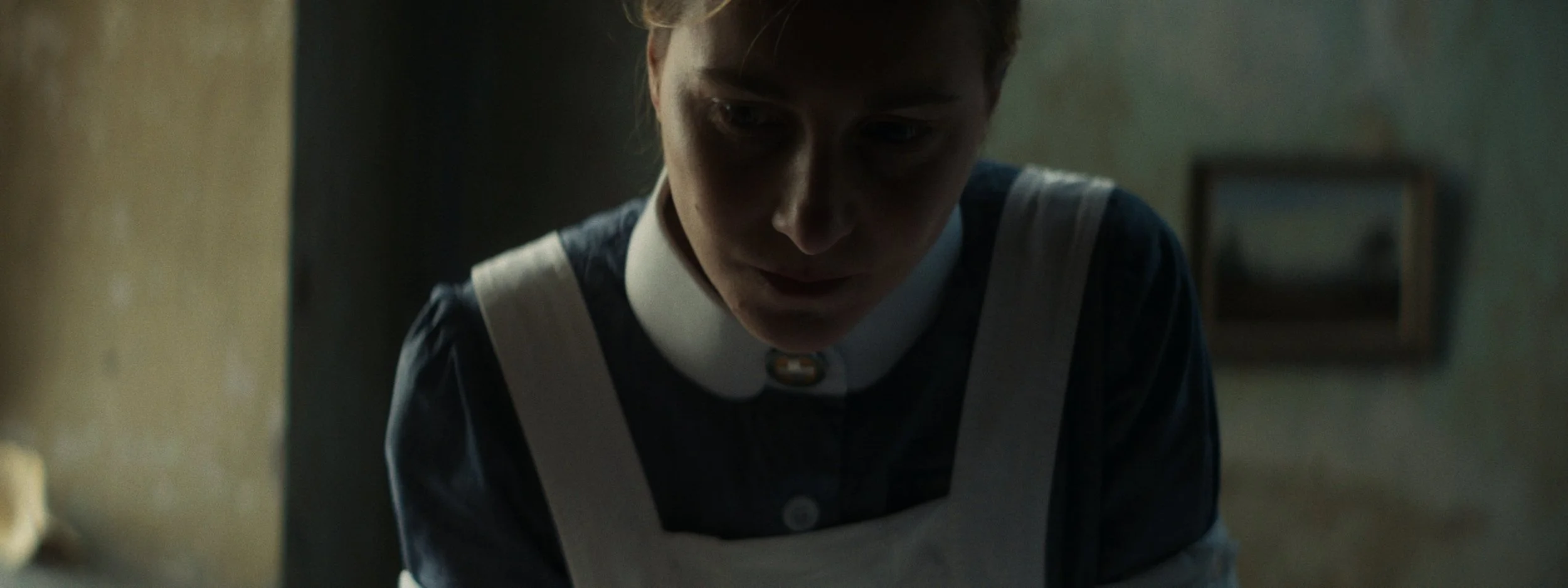A young woman with short hair wearing a dark uniform and white apron looking down thoughtfully in a dimly lit room.
