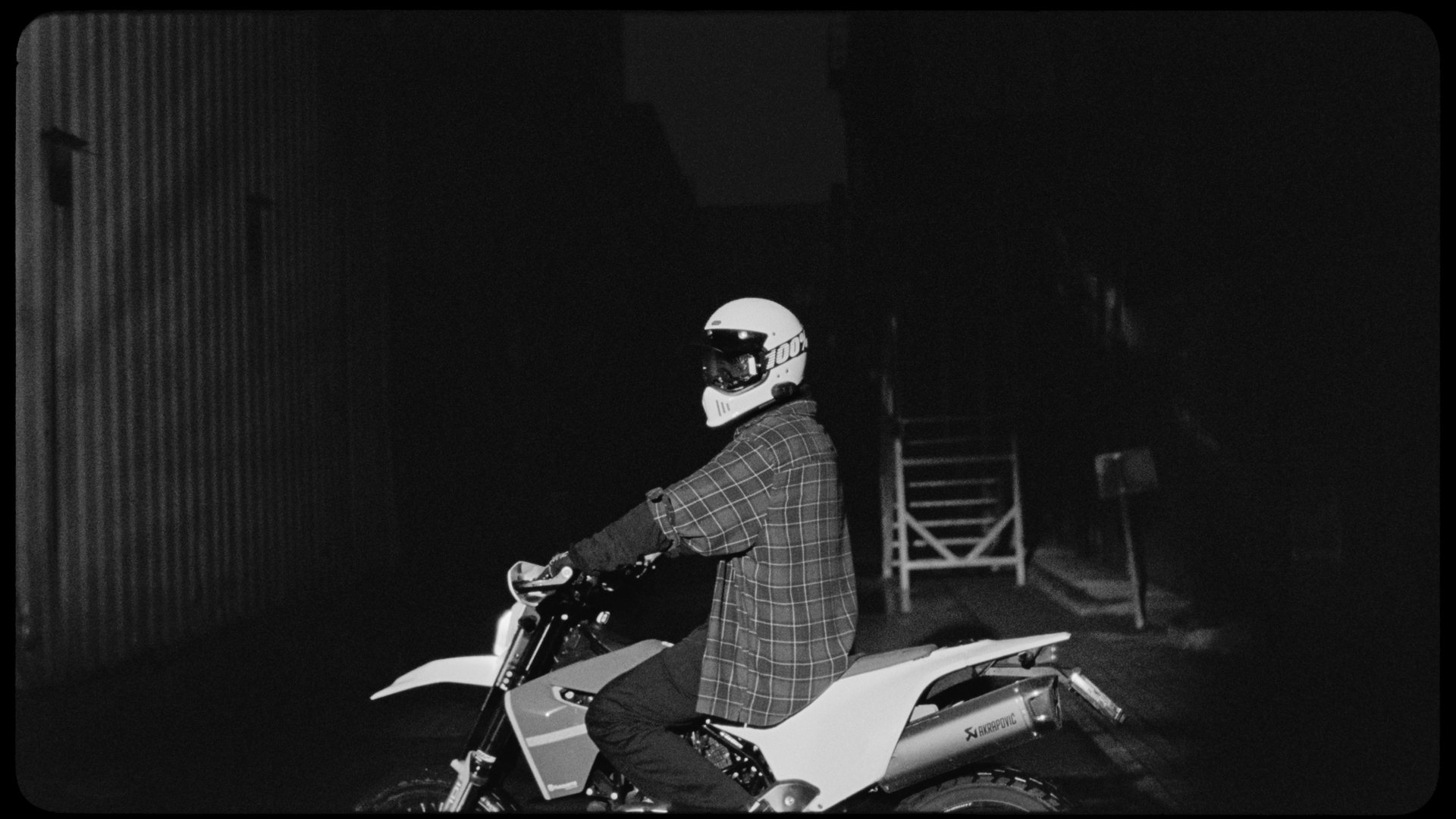 A person riding a off-road motorcycle at night, wearing a white full-face helmet and a plaid shirt, with a dark background and some outdoor furniture nearby.
