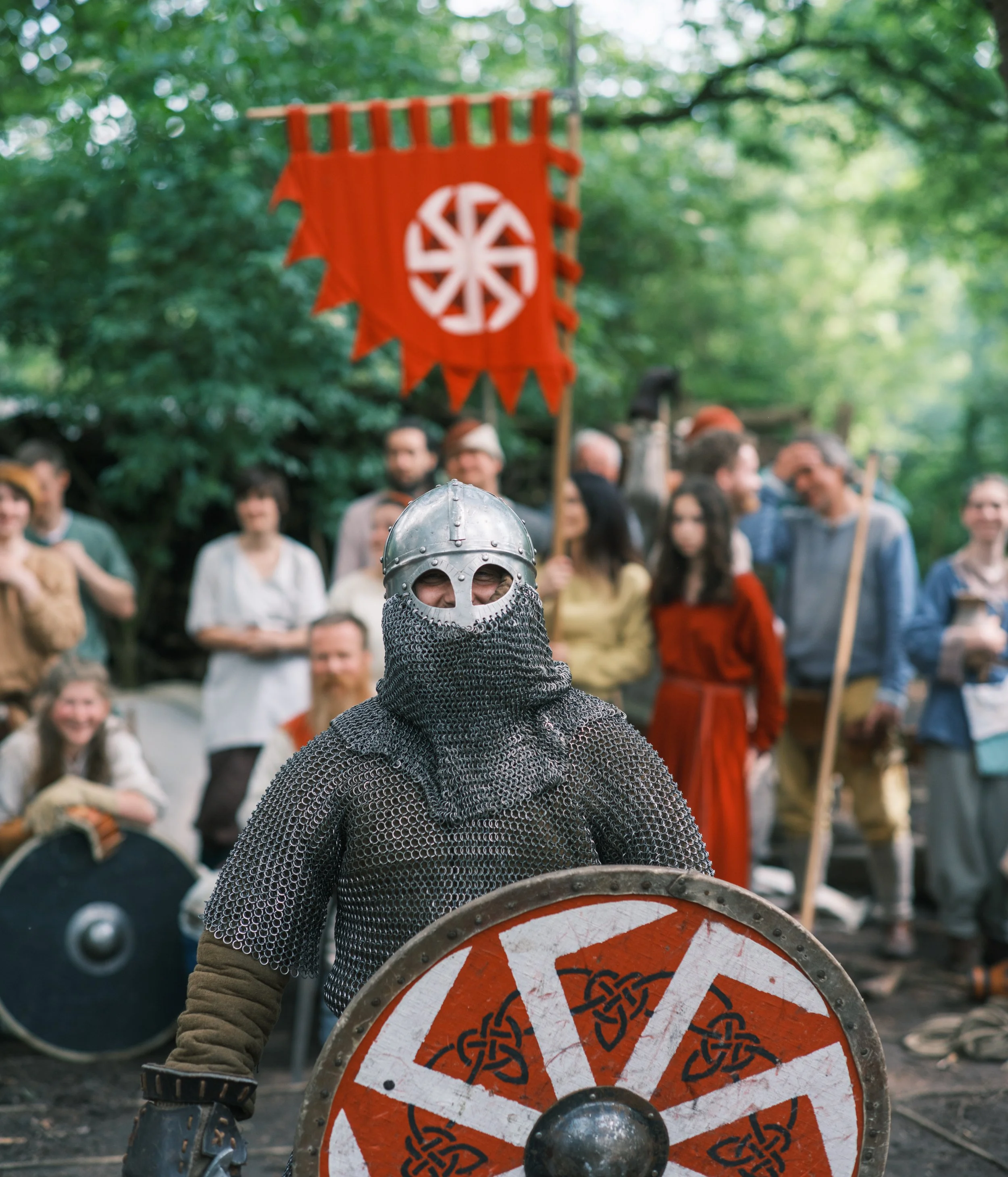 Person dressed as a medieval knight wearing chainmail and a helmet, holding a round shield with Viking knot patterns, standing outdoors at a historical reenactment event with a crowd and a red banner in the background.