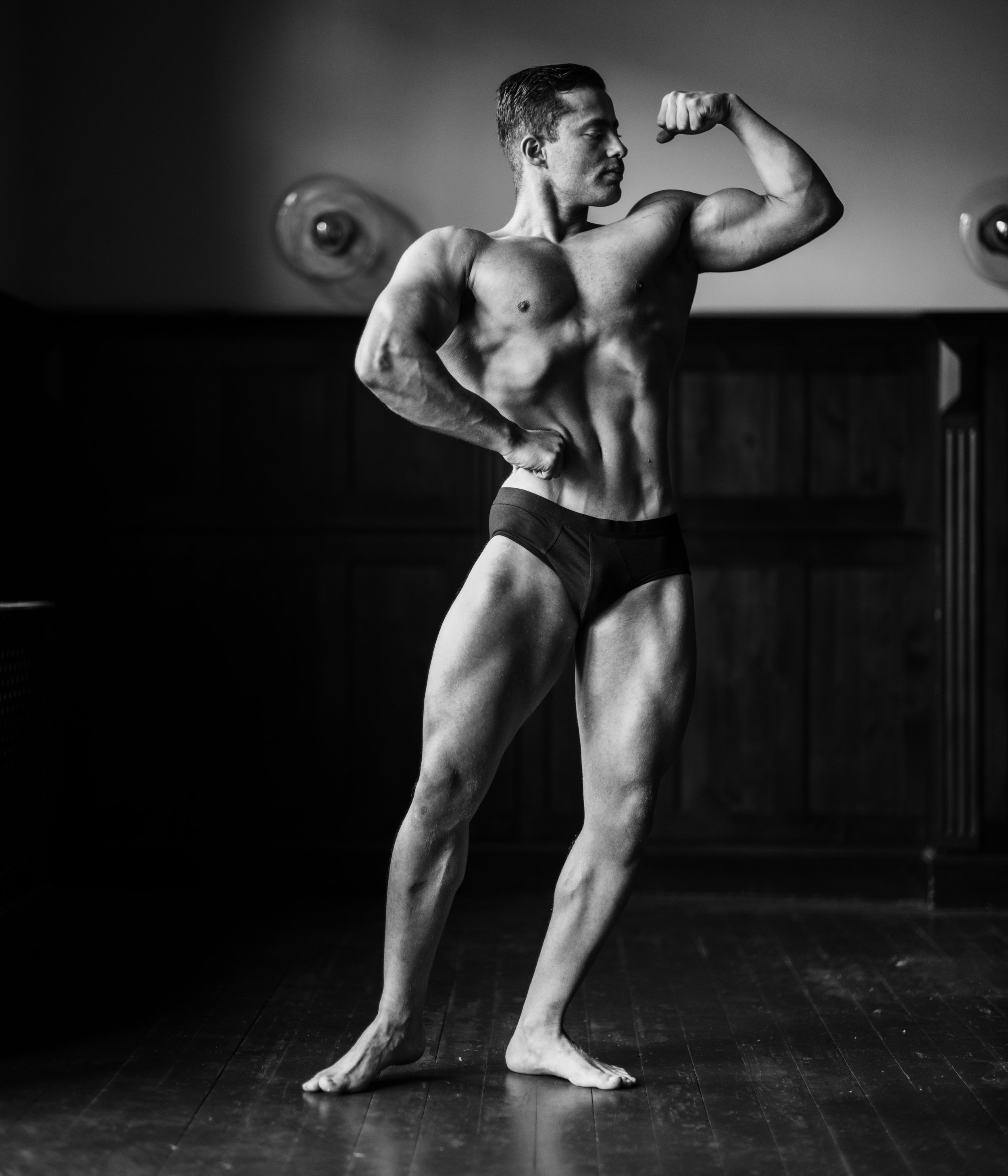 Black and white photo of a muscular man posing shirtless, flexing his right arm, with one hand on his hip, standing on a wooden floor, against a plain background.