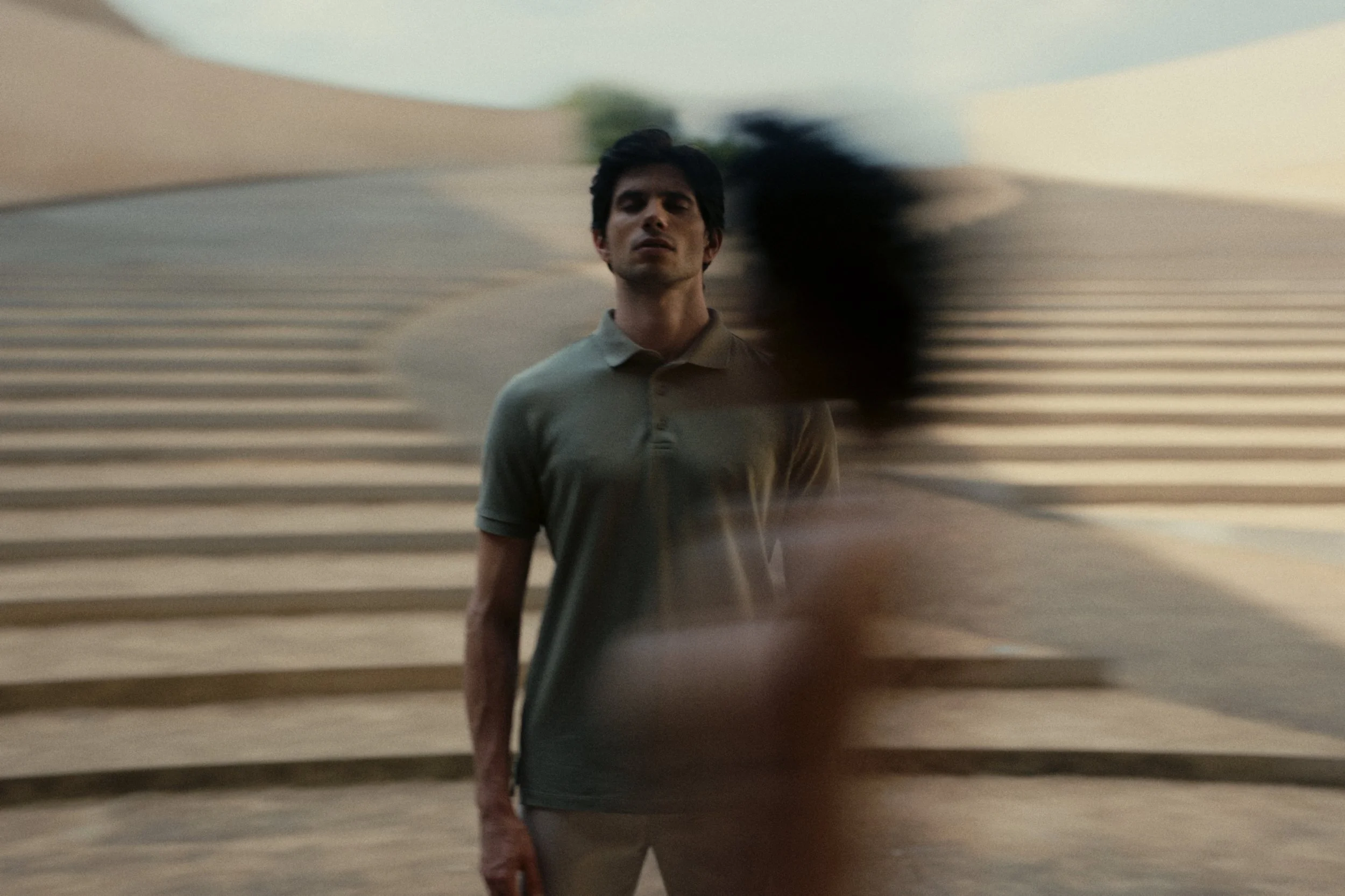 A man in a beige polo shirt standing in front of a wall with horizontal stripes, with blurred motion affecting part of the image.