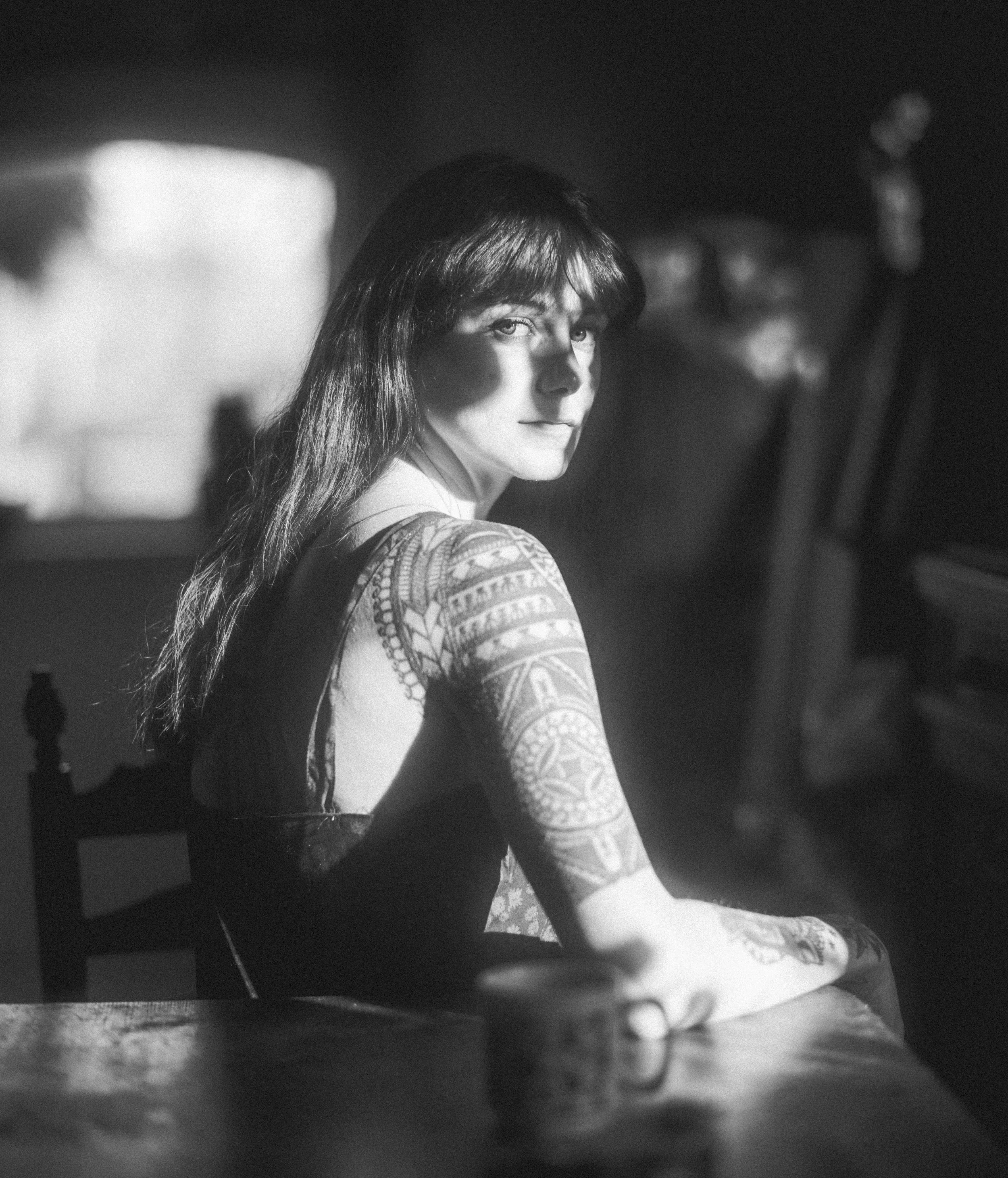 A black and white photograph of a young woman with long hair and tattoos, gazing over her shoulder while seated in a dimly lit environment.