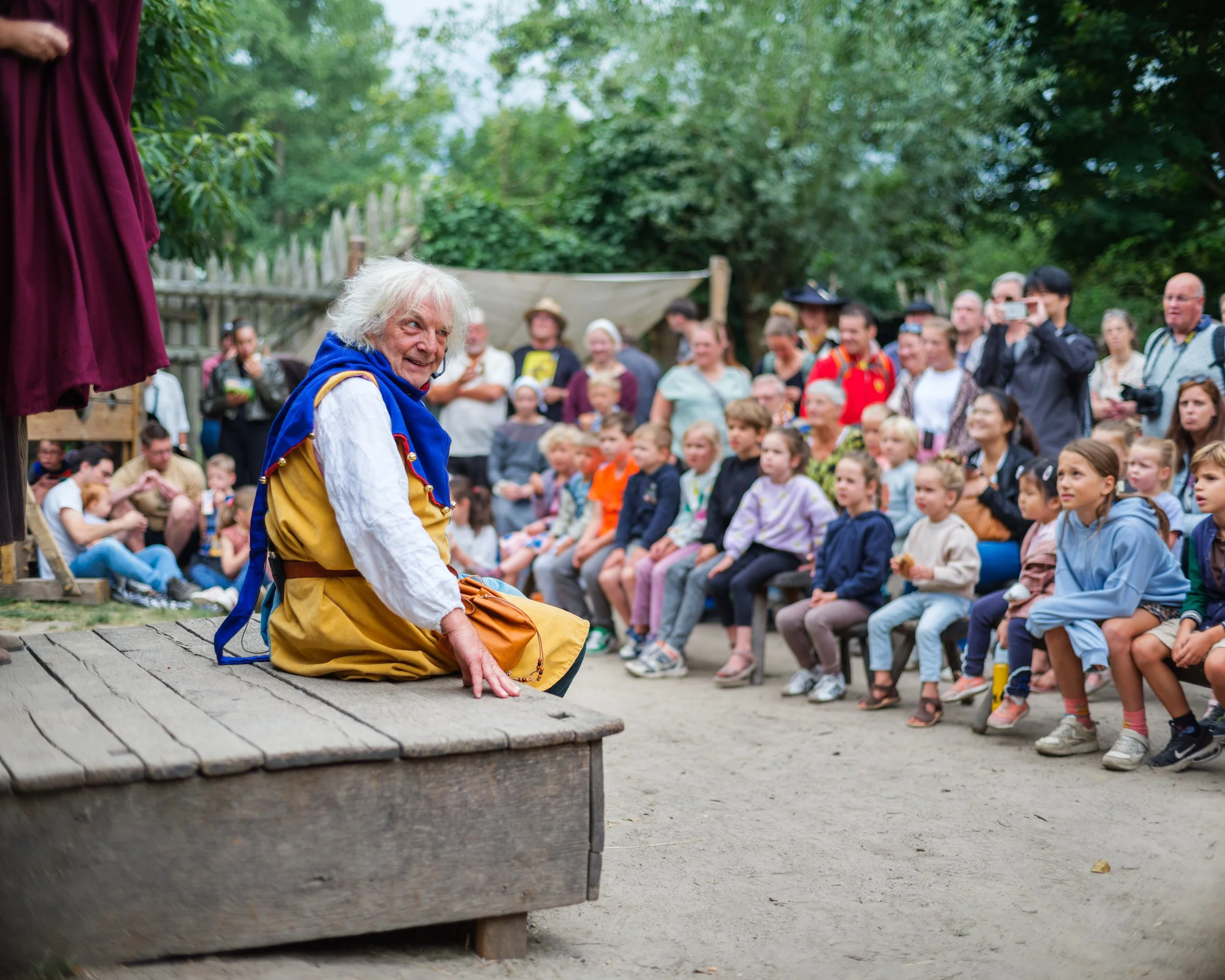 A person dressed in medieval costume kneeling on stage with an audience of children and adults watching outdoors, surrounded by trees.