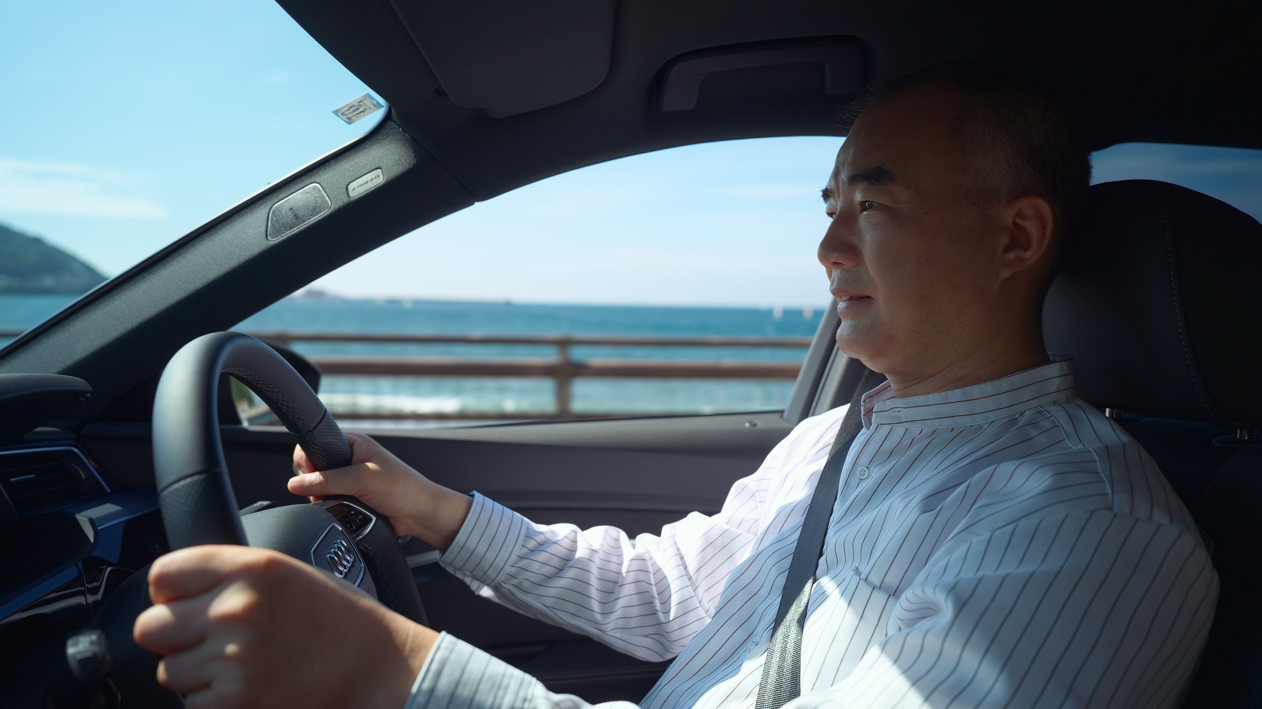 A man driving an Audi car along a coastal road with the ocean and a mountain visible in the background.