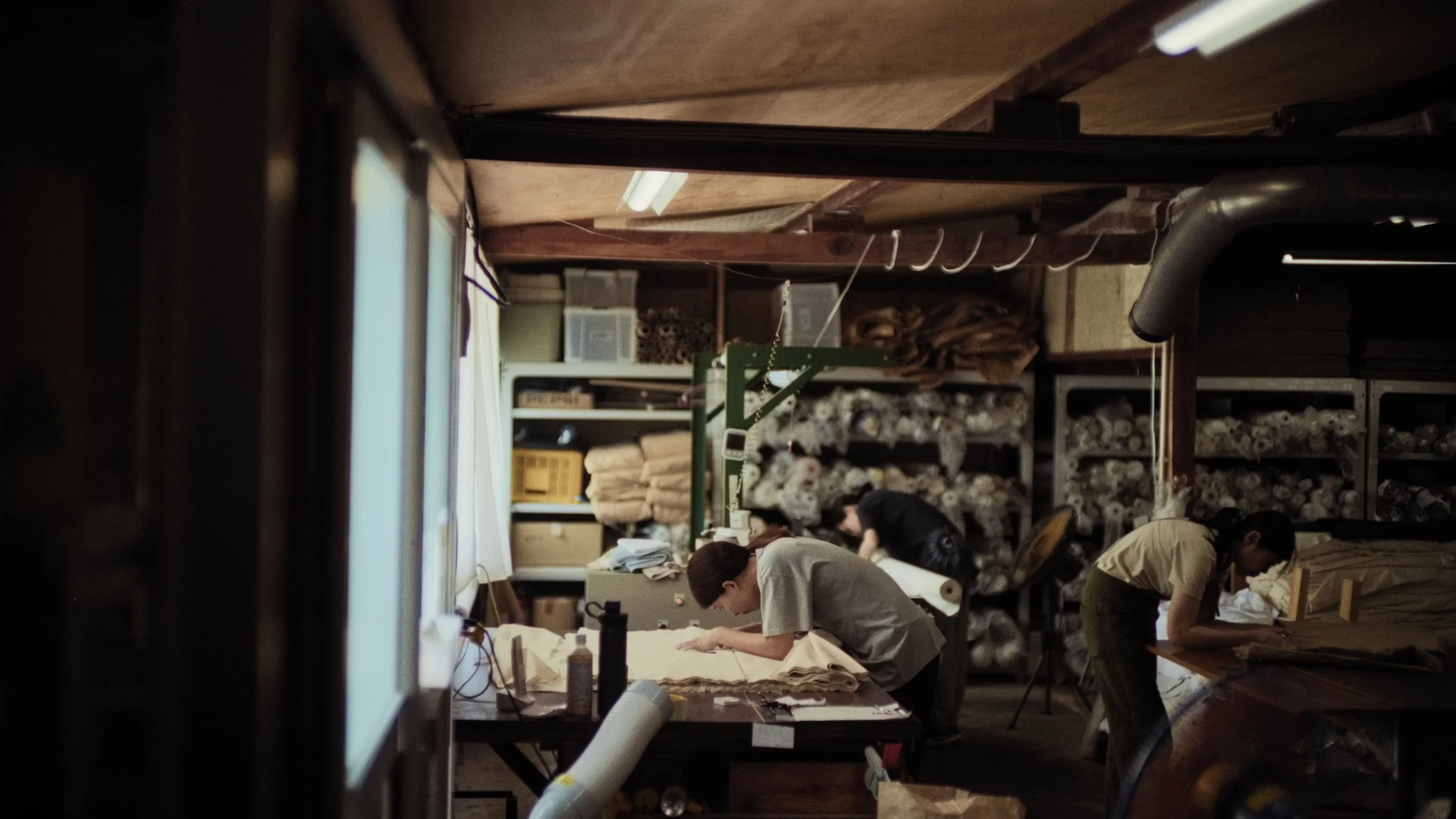 People working on craft projects in a cluttered workshop with shelves filled with fabric and supplies.