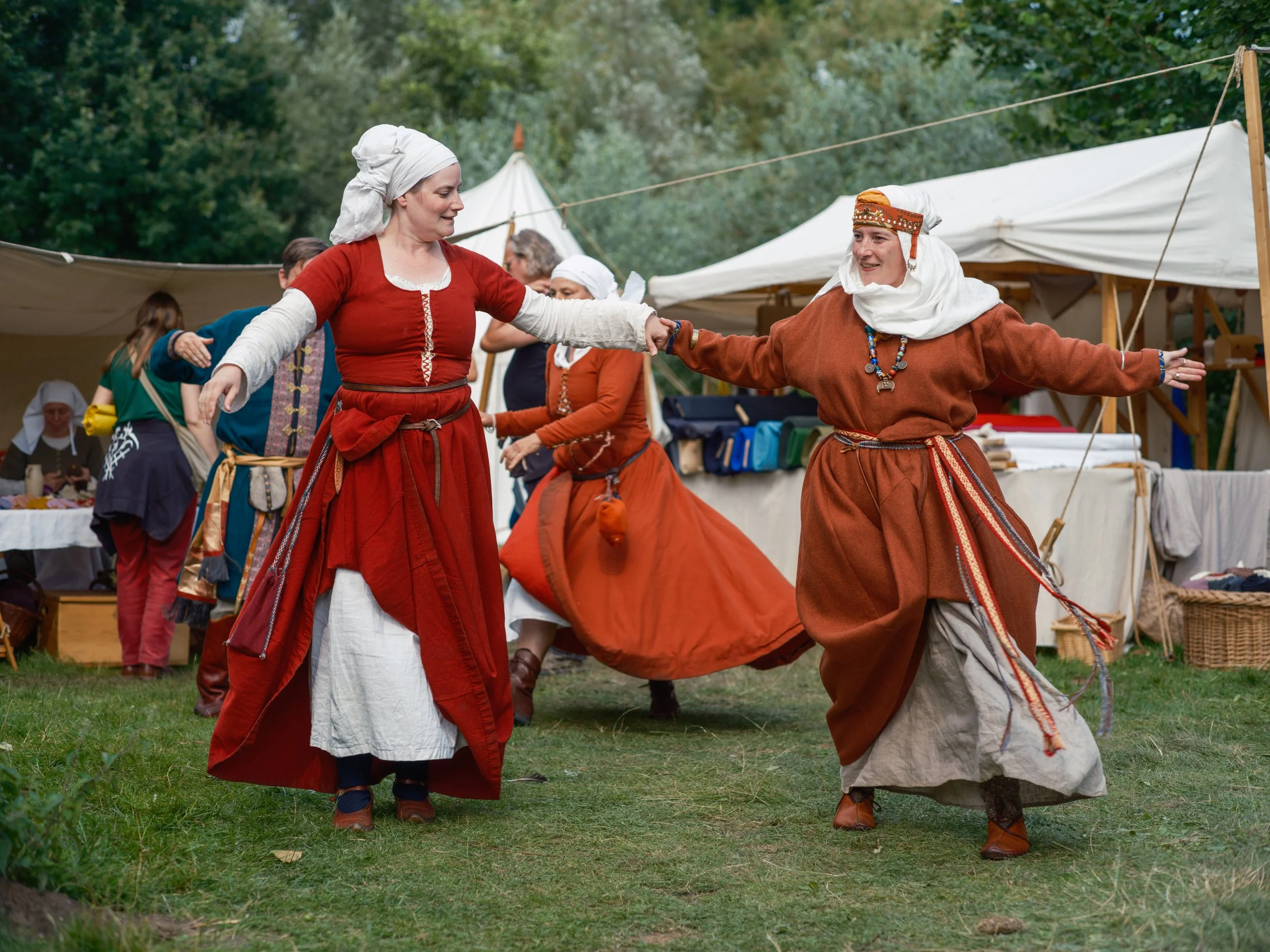 Two women dressed in medieval-style clothing dancing at an outdoor event with tents and trees in the background.