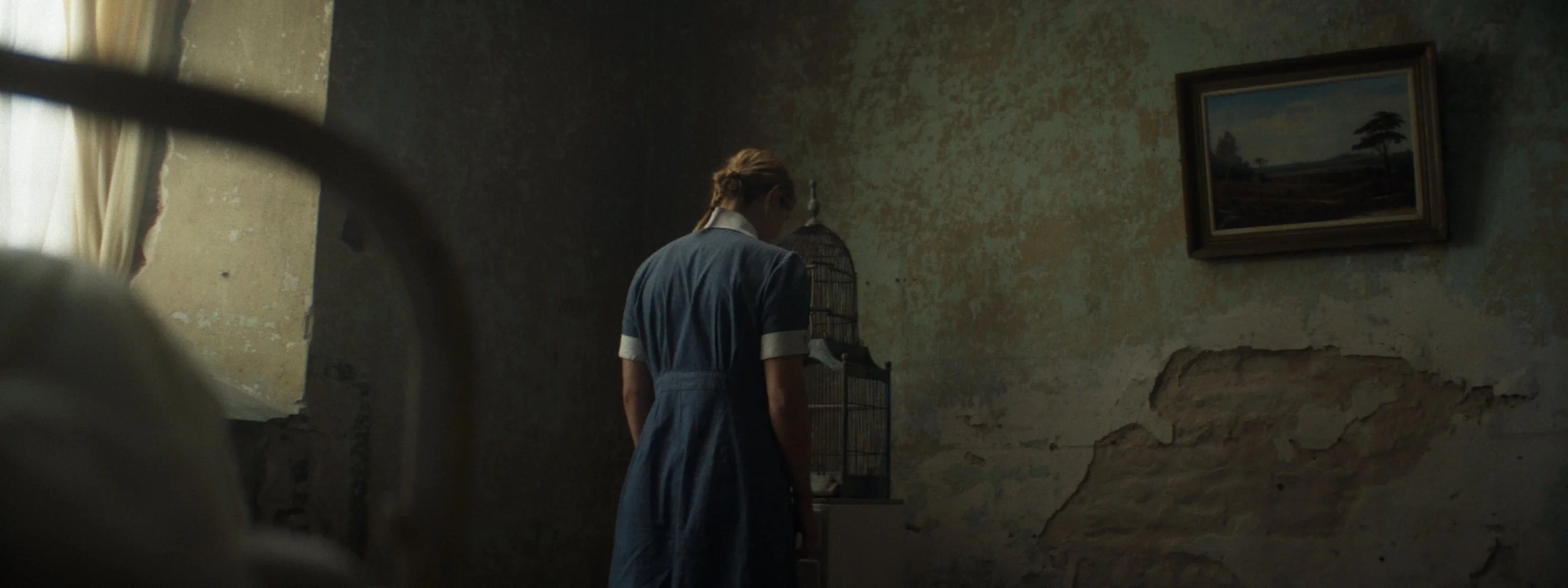 A young woman in a vintage blue dress with white collar and cuffs standing in a dimly lit, worn-down room with peeling paint on the wall, looking down. There is a framed painting of a landscape hanging on the wall and a birdcage on a small table.