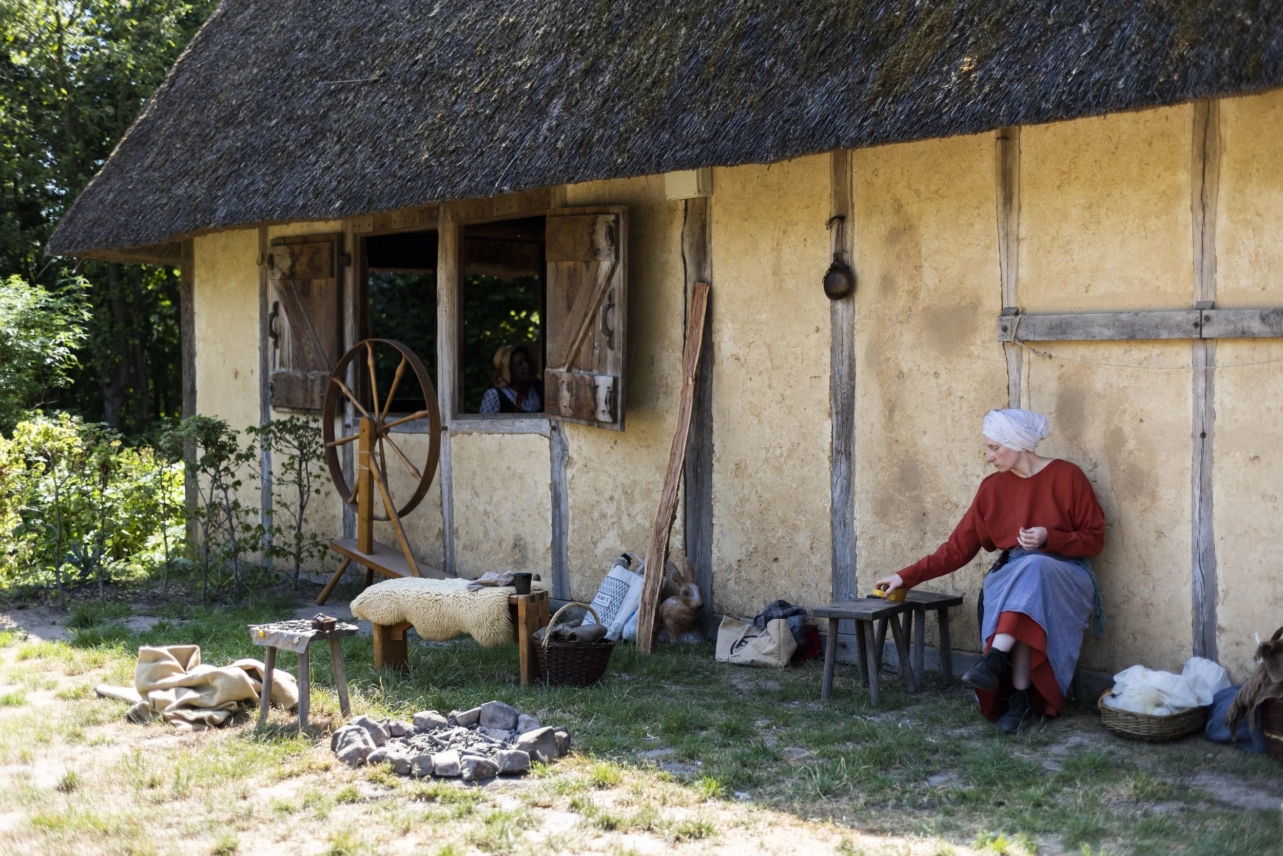 A woman in a red dress and white headscarf sitting outside a thatched-roof cottage, engaging in some activity with her hands, surrounded by various items and animals.