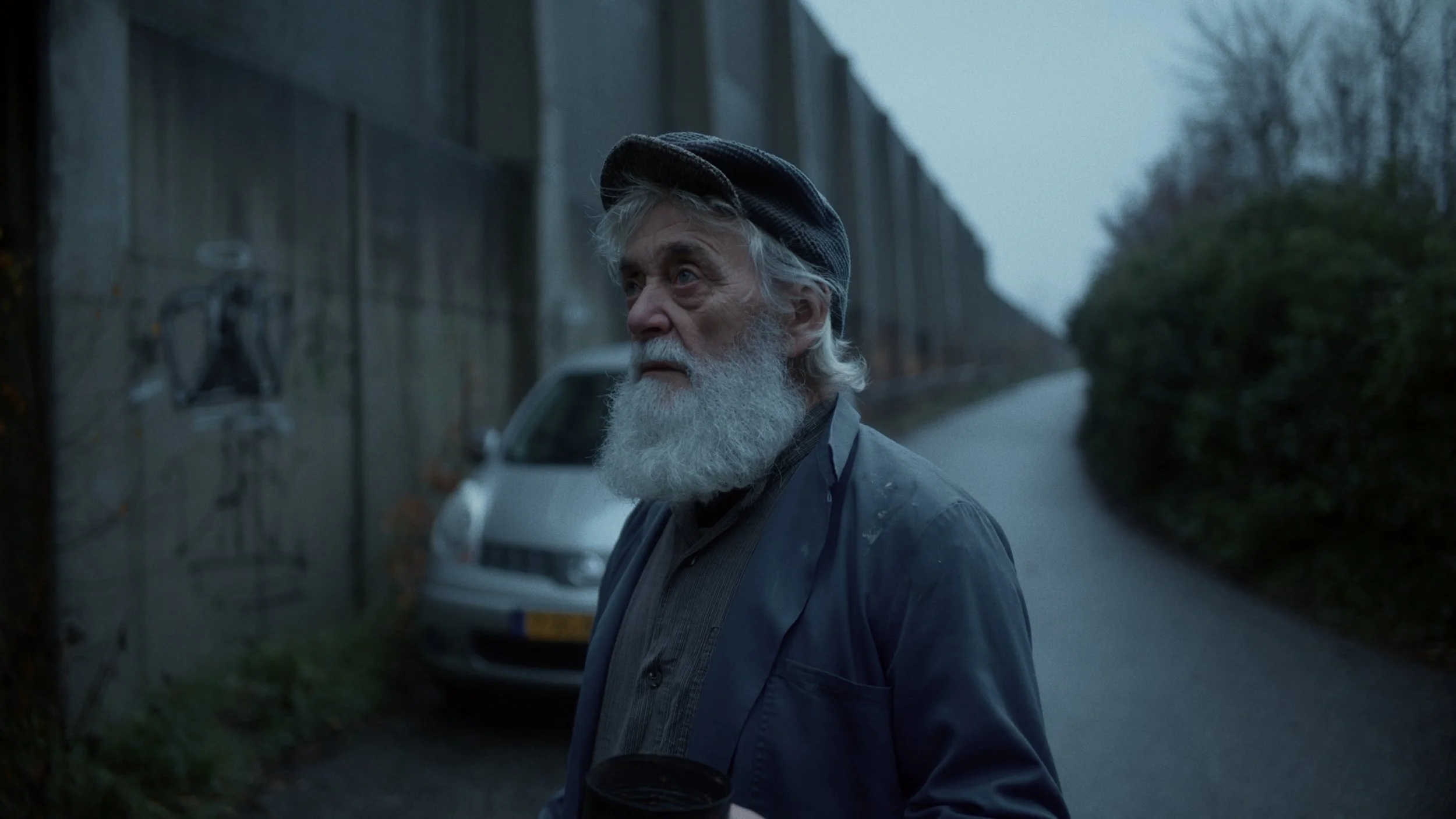 An elderly man with a white beard and gray hair, wearing a dark cap and jacket, stands outdoors on a rainy day near a gray wall and parked car, with trees and a road in the background.