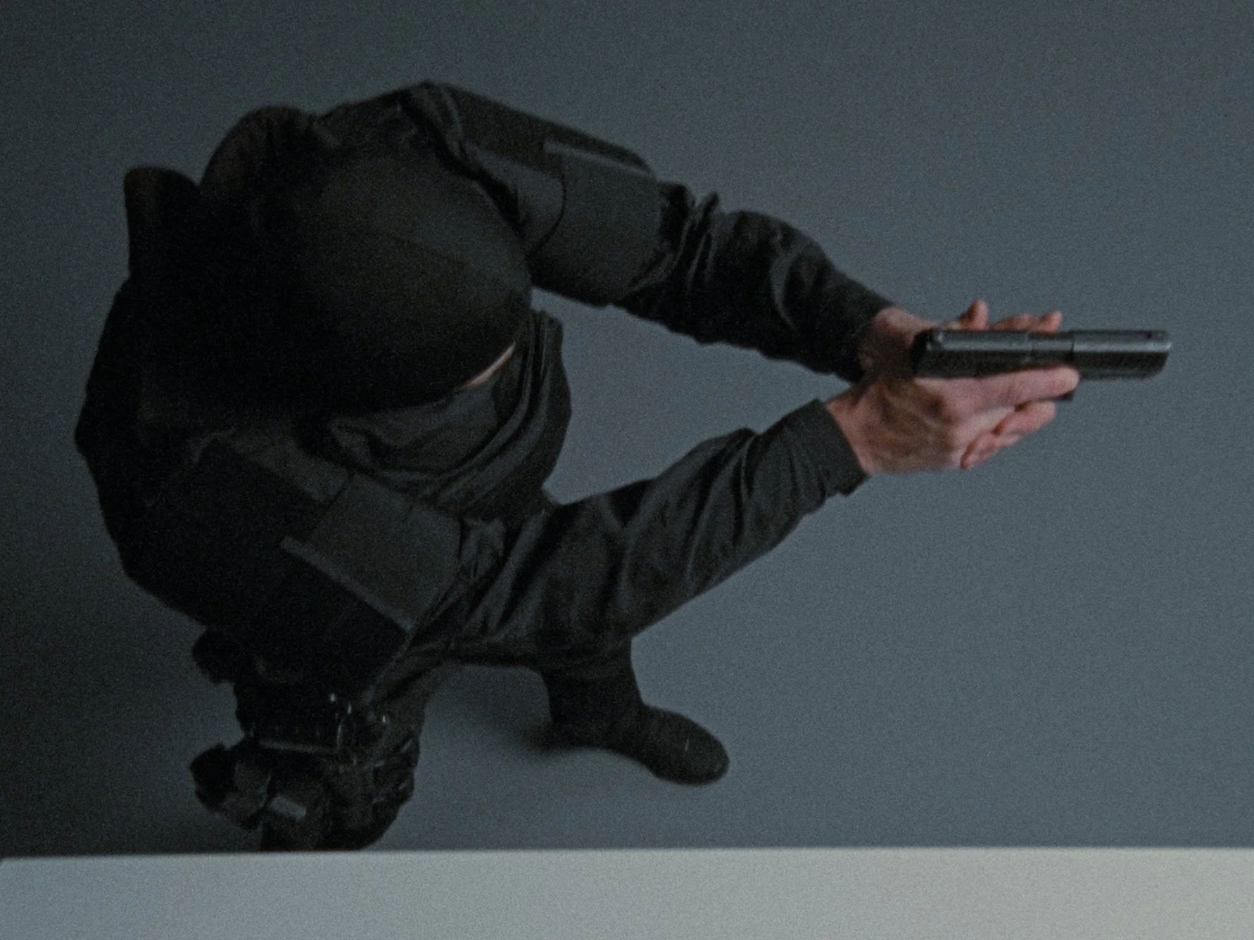 A person dressed in black tactical clothing, crouching and holding a handgun with both hands, aiming it downward against a gray wall.