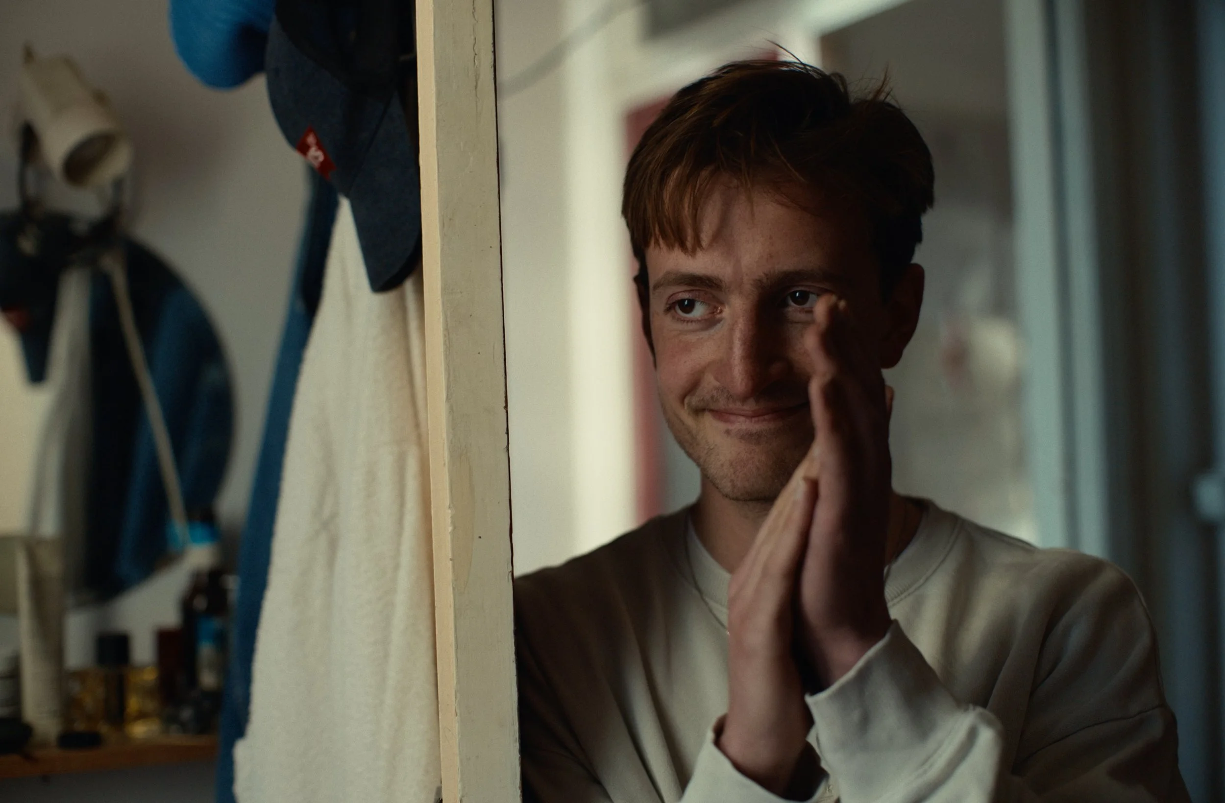 A young man with brown hair and a light brown shirt presses his palms together in a prayer or contemplative gesture, peering into a mirror in a room with bookshelves and hanging clothes.