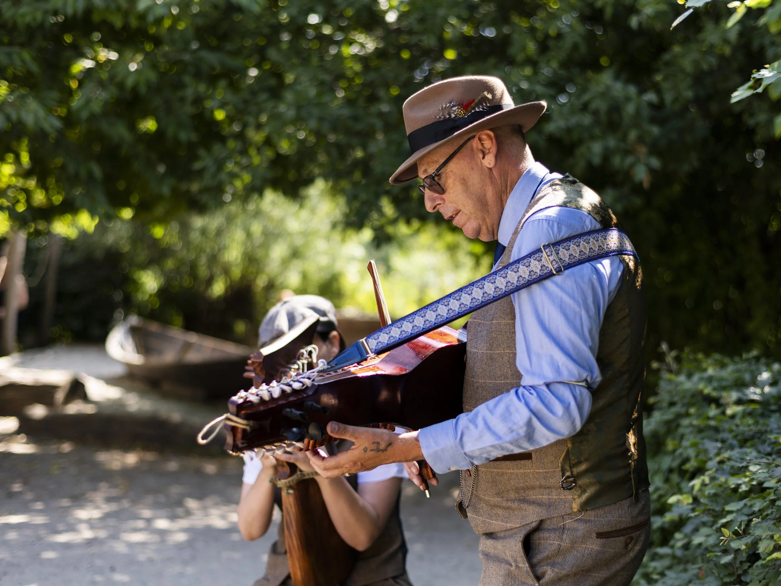 A man wearing a beige hat with a feather, black glasses, a blue shirt, a plaid vest, and beige pants is playing a guitar outdoors, with other people and trees in the background.
