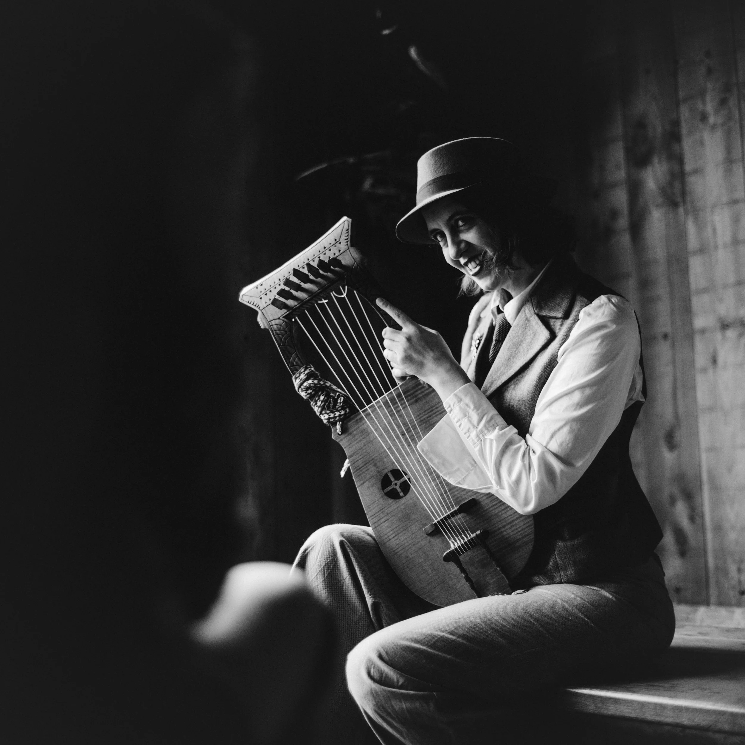 Black and white photo of a person wearing a top hat and collared shirt, smiling while playing an acoustic guitar. The person is looking to their right and the background is dark.