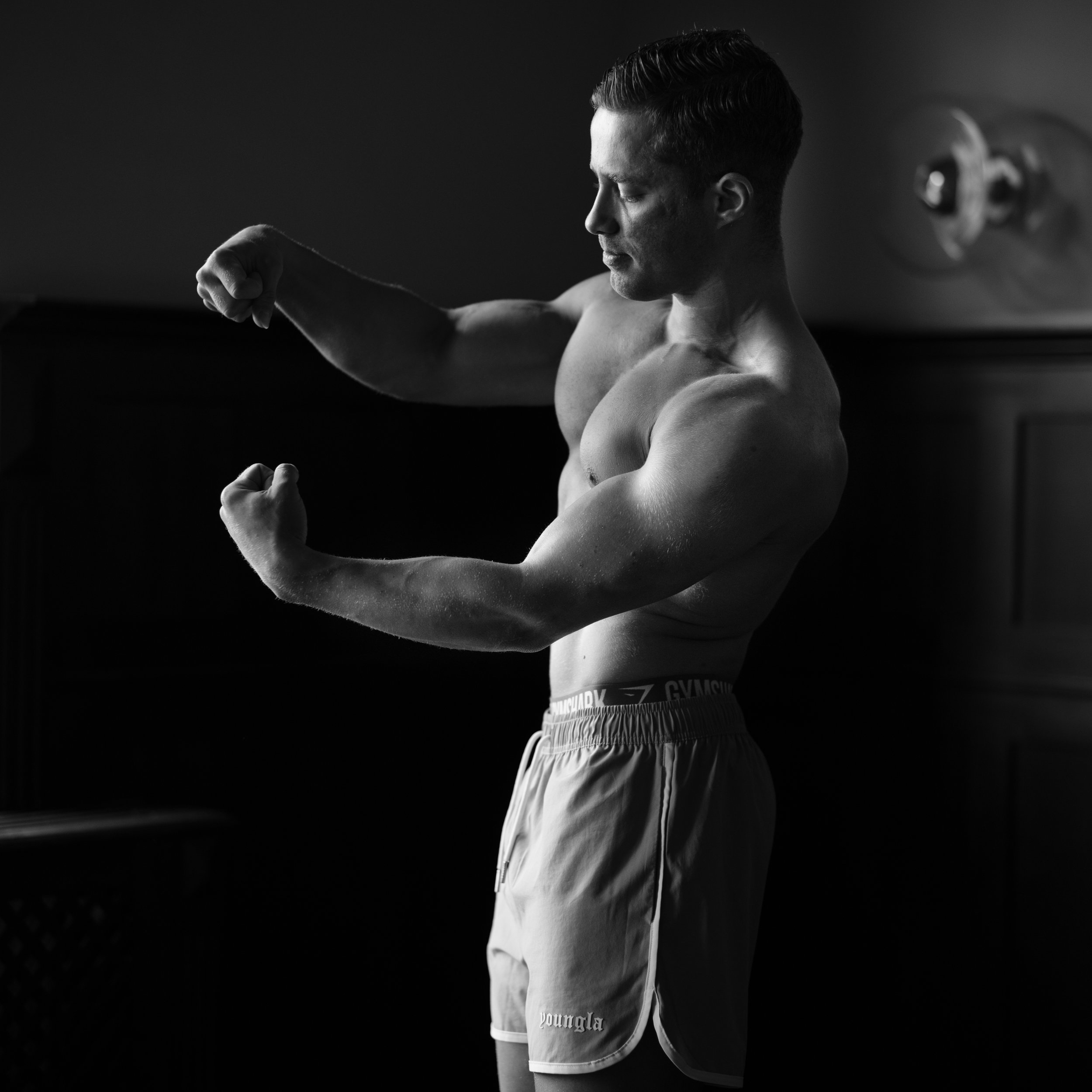 Black and white photo of a shirtless young man with slicked-back hair, flexing his arm in a gym or training environment, with a wall-mounted discus plate visible in the background.