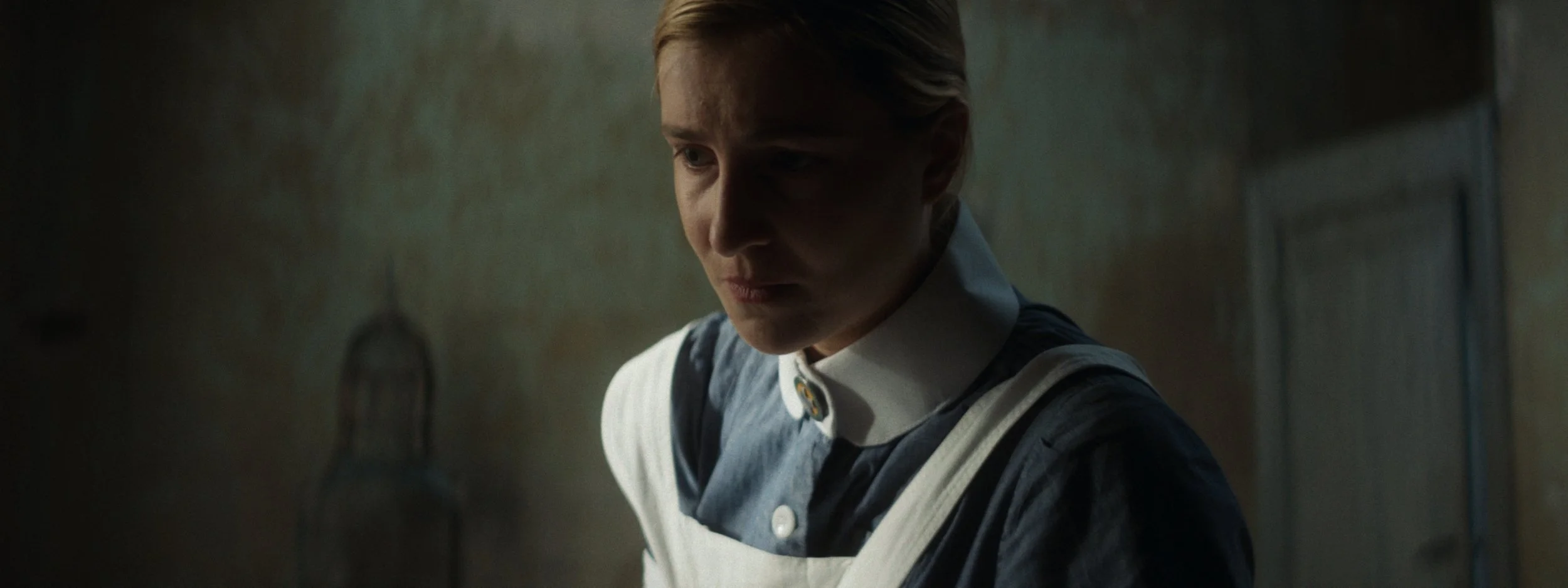 A young woman with red hair, wearing a navy blue uniform and a white apron, looks down thoughtfully in a dimly lit kitchen.
