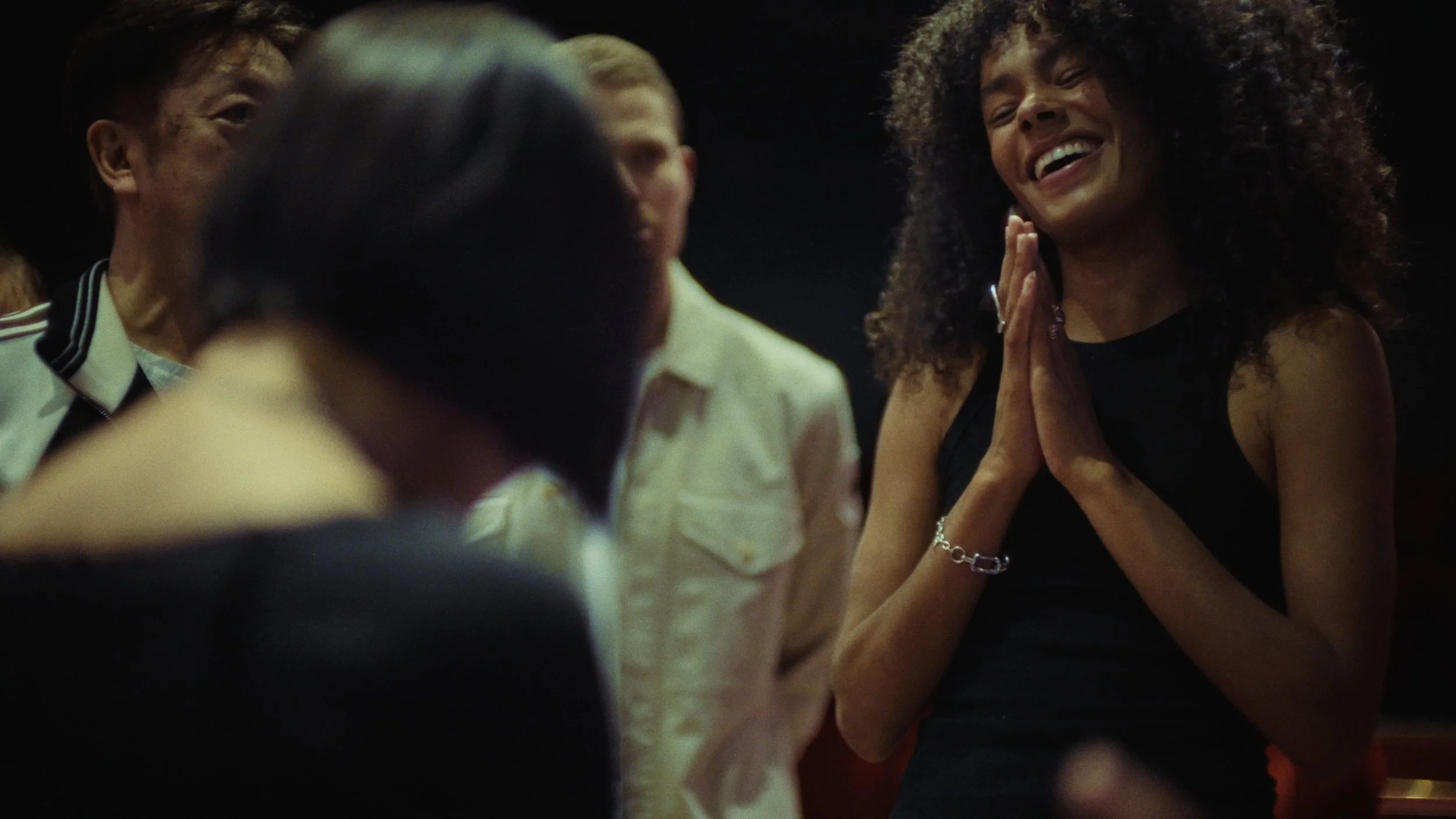 A woman with curly hair smiling and clasping her hands together, surrounded by other people in a dimly lit setting.