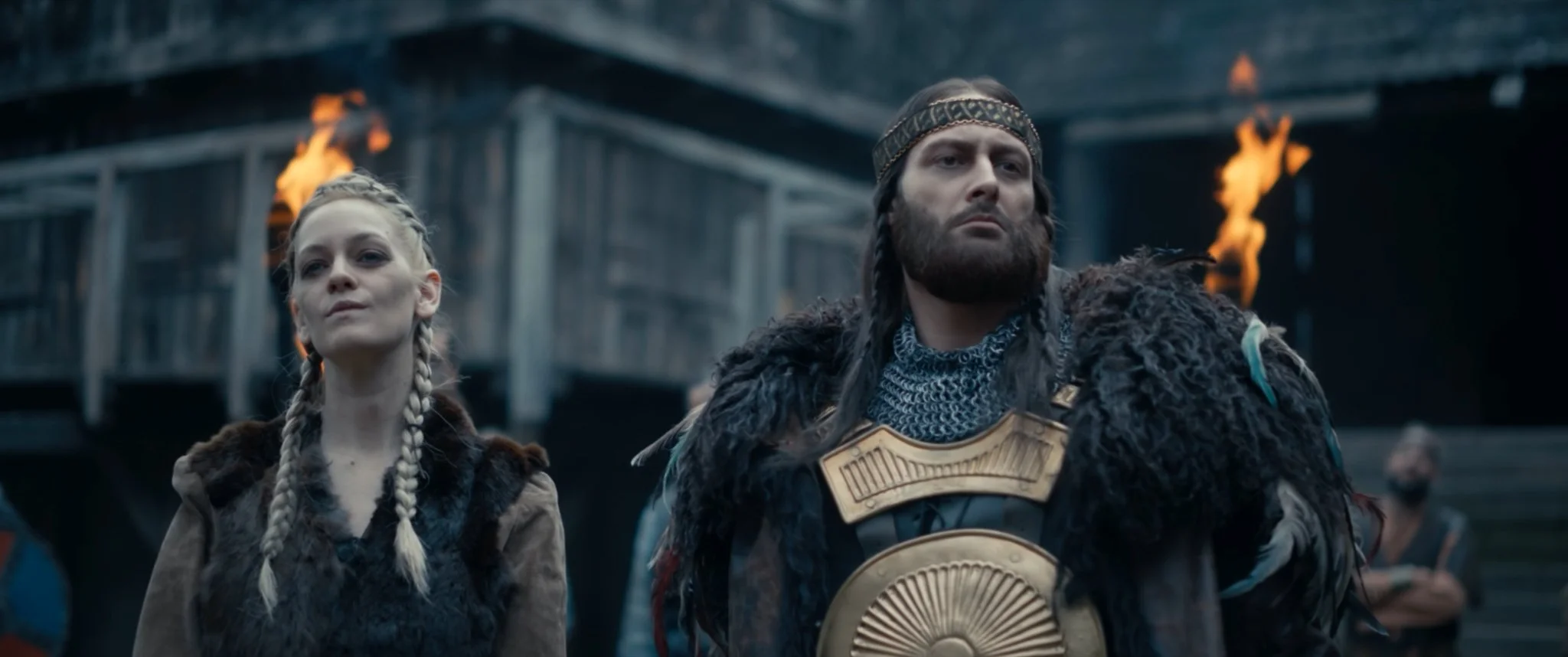 Two people wearing medieval Viking costumes, standing side by side outdoors, with wooden structures and fire behind them.