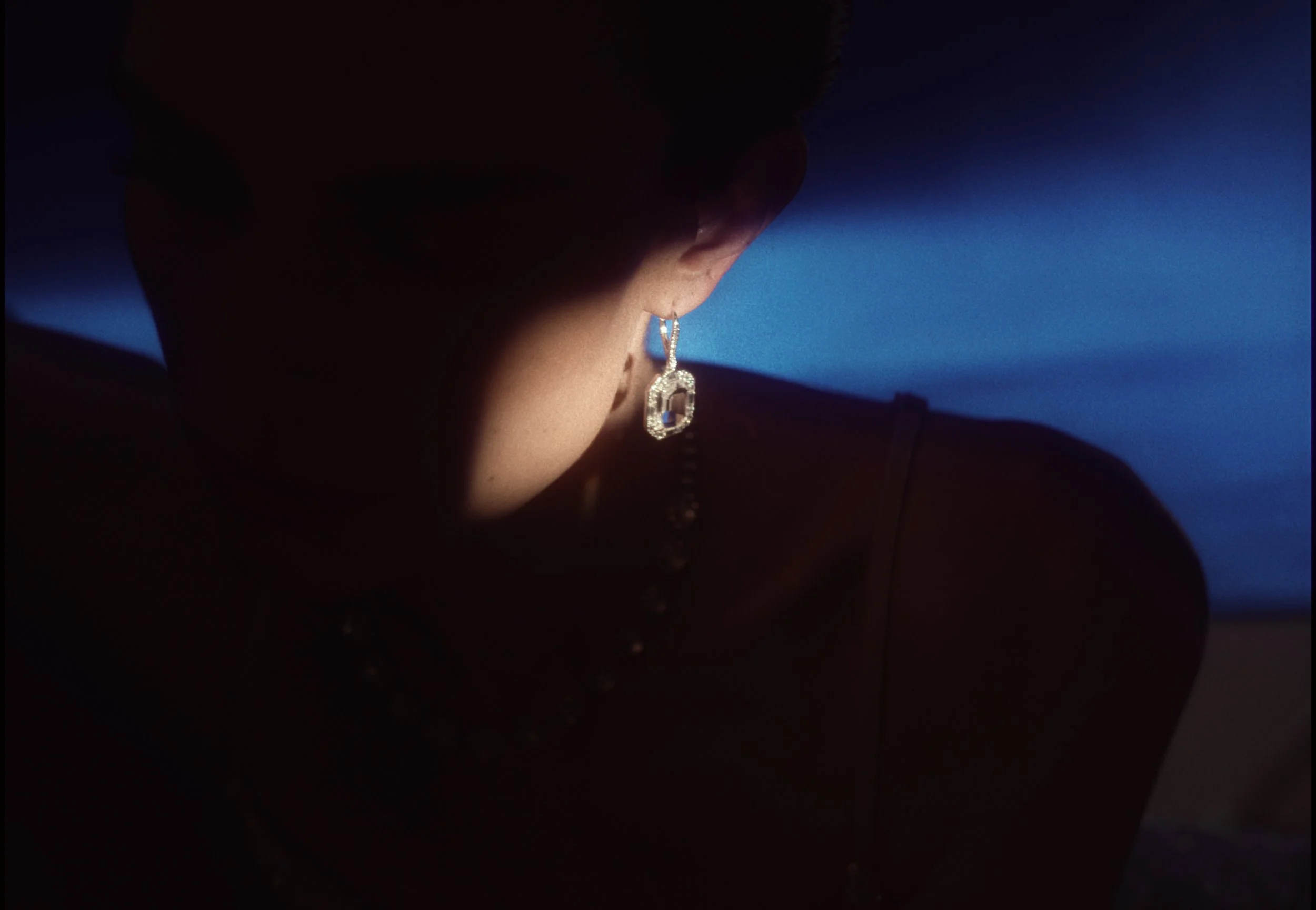 Close-up of a woman wearing a diamond earring, with her face partially obscured by darkness and a blue background.
