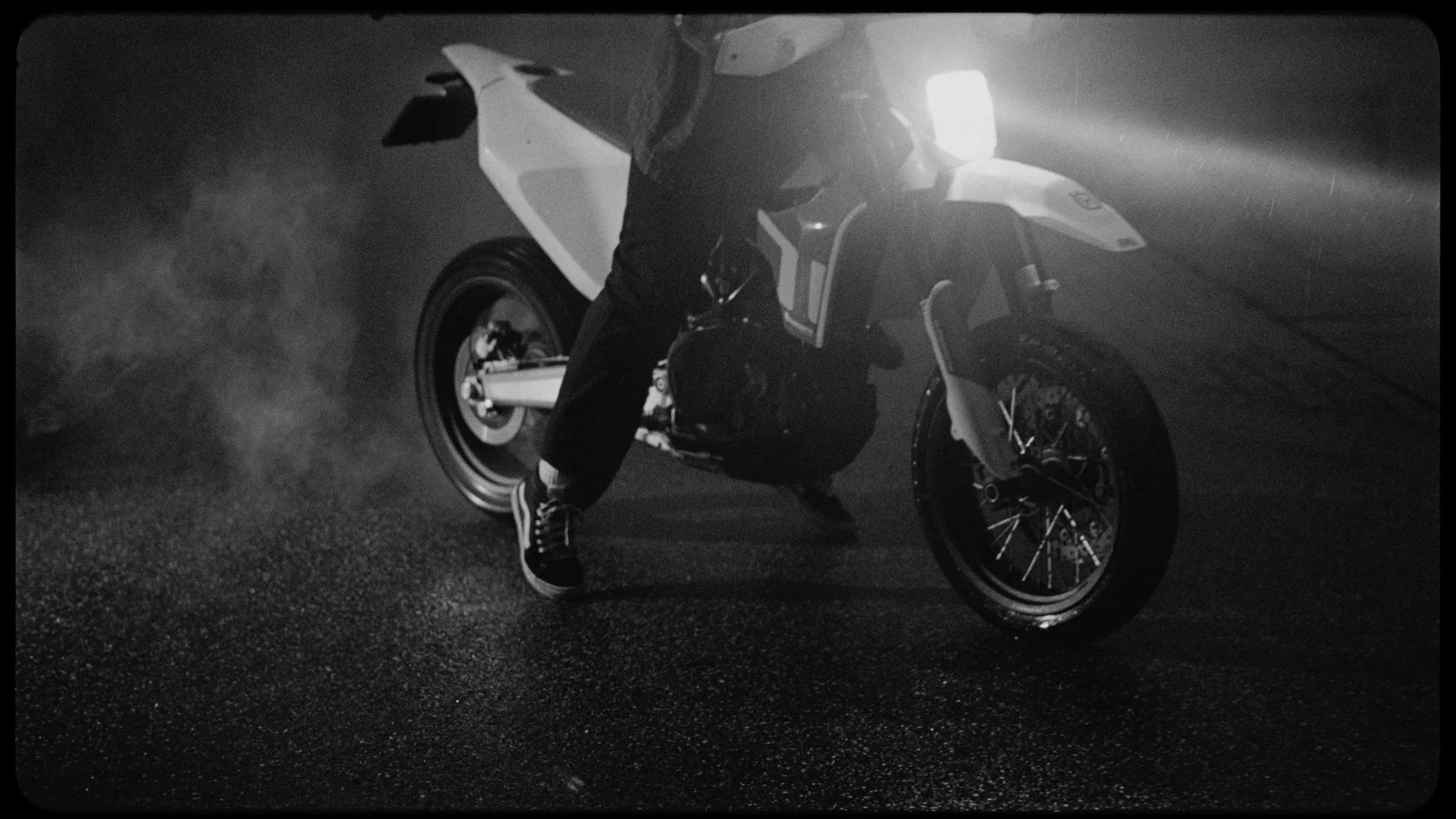 Person wearing sneakers doing a wheelie on a motorcycle at night, with headlights on and smoke or dust in the air, in black and white.