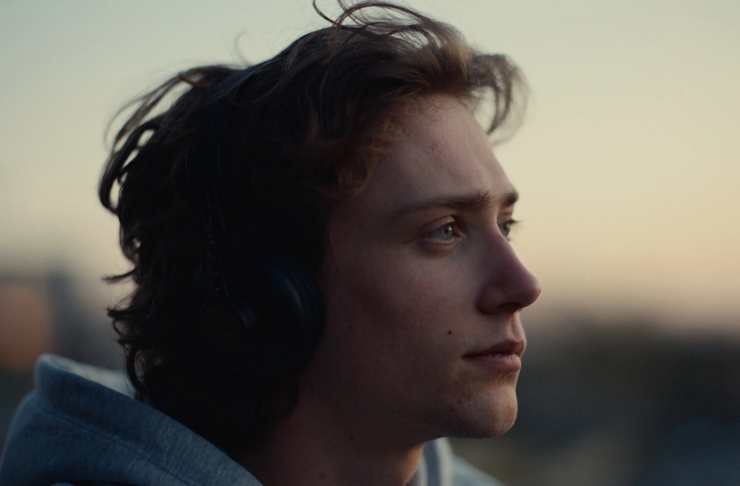 Close-up of a young man with curly dark brown hair and blue eyes, looking thoughtfully into the distance under natural lighting.