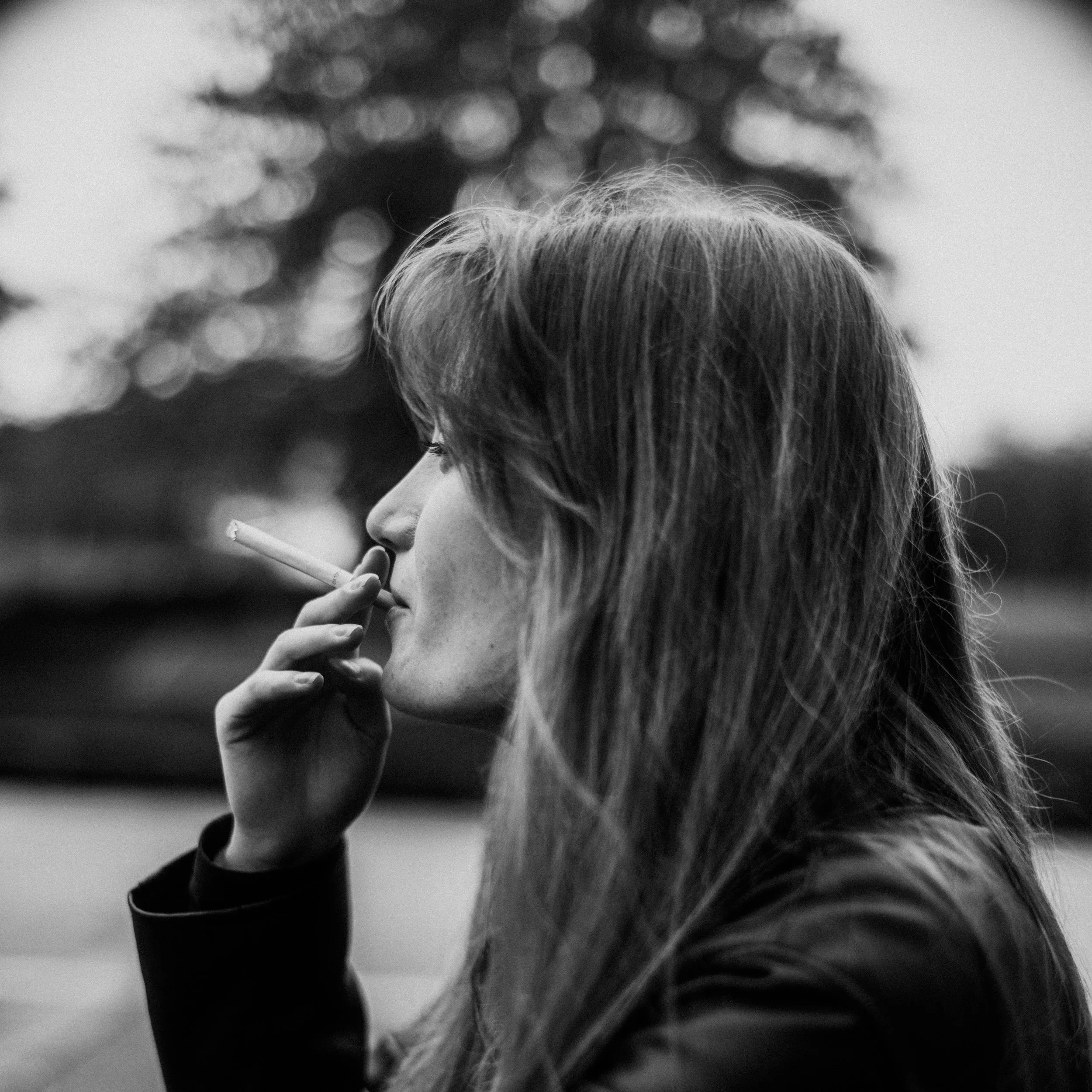 A person with light-colored hair smoking a cigarette outdoors, with blurred trees in the background, black and white photograph.