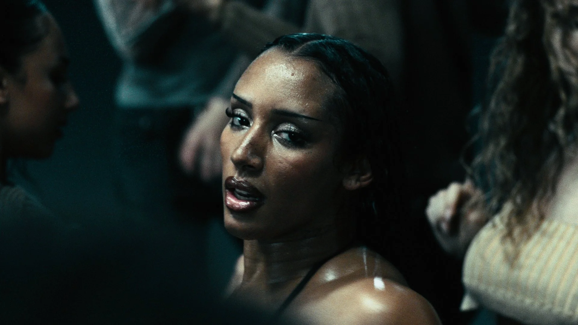 Close-up of a woman with wet hair and makeup, surrounded by others in a dimly lit setting, with a dark background.