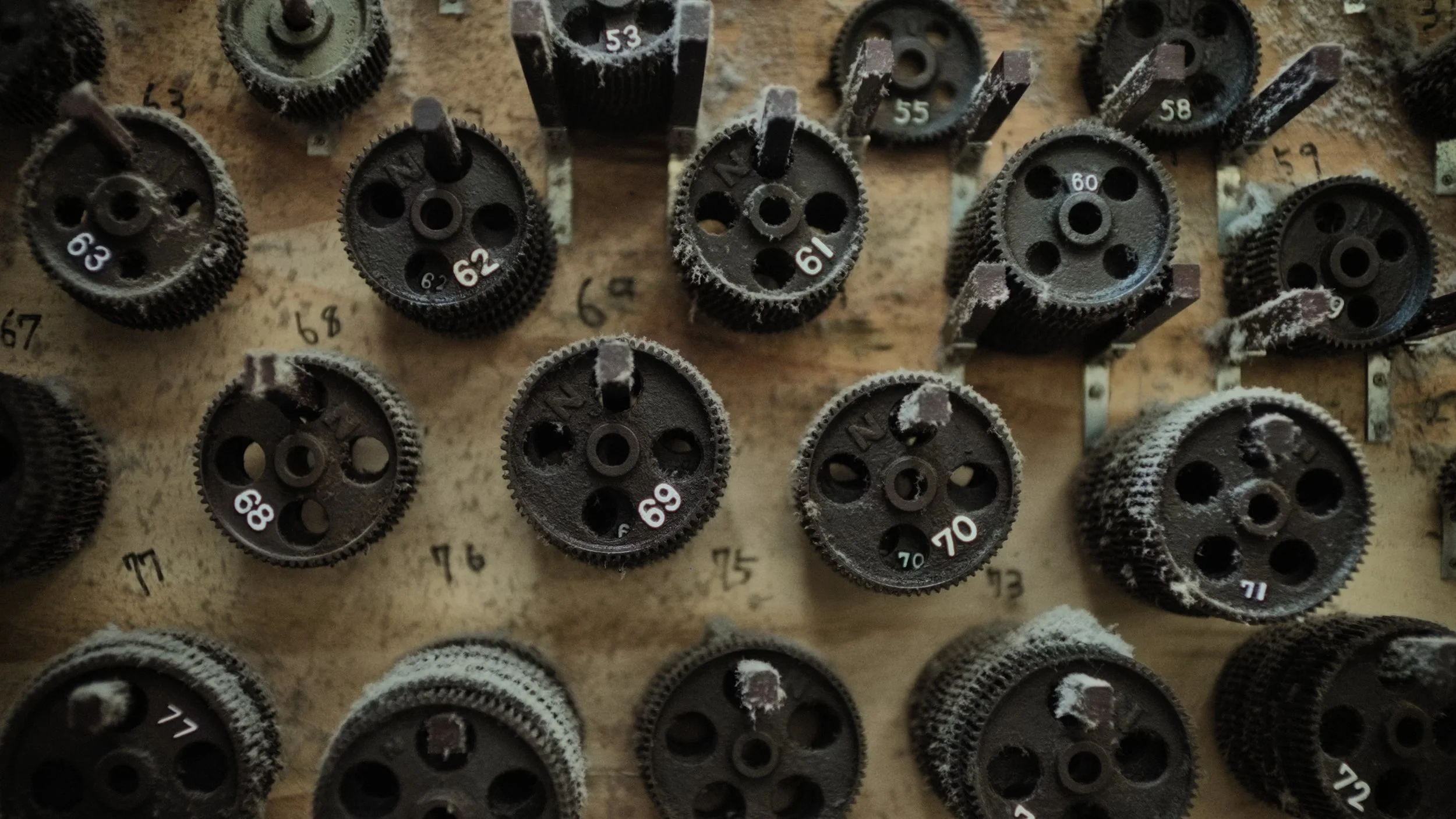 Close-up of dusty black gears with numbered white labels on a wooden surface, arranged in rows.