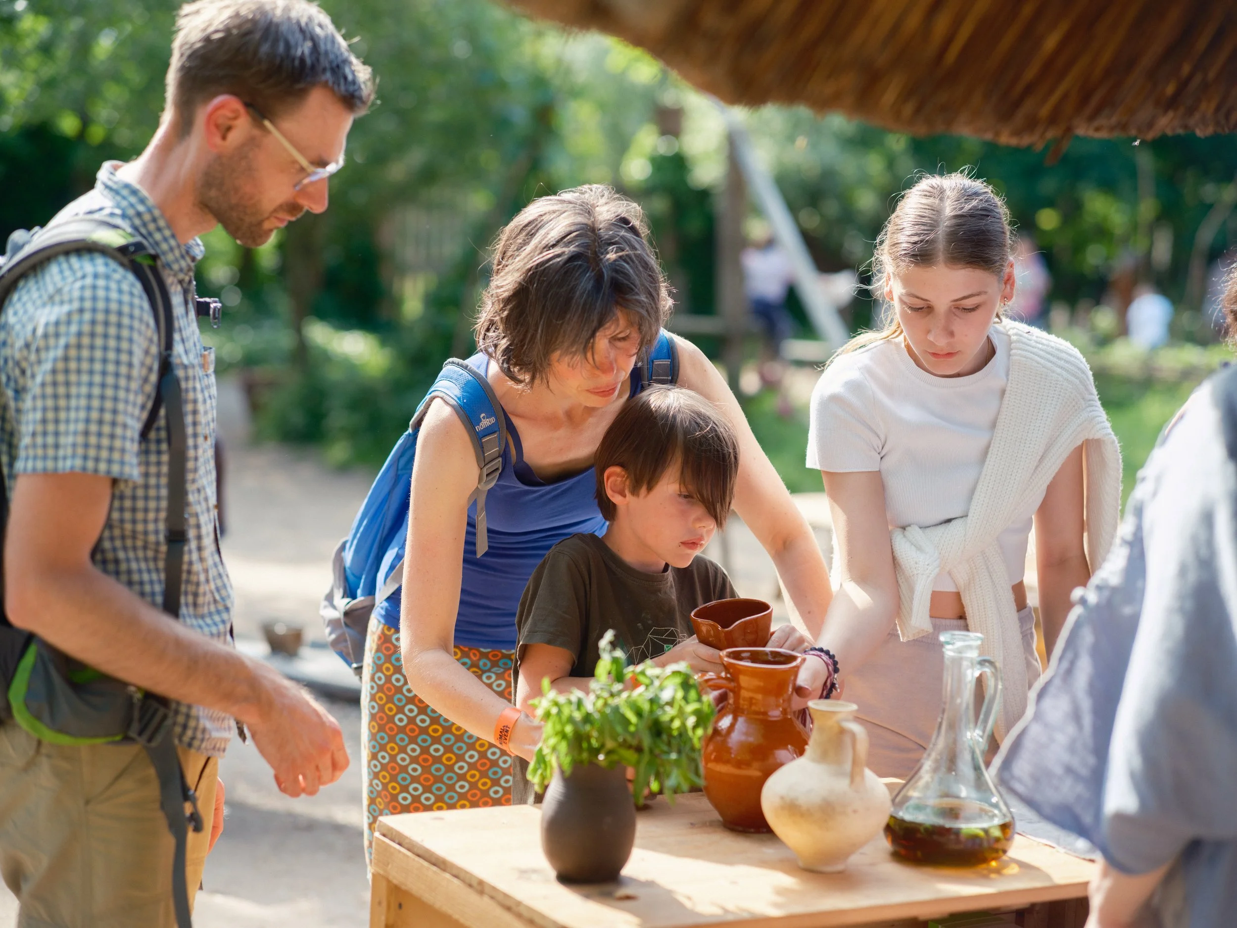 People gather outdoors around a table displaying pottery and glassware, engaging in a pottery or craft activity with trees in the background.