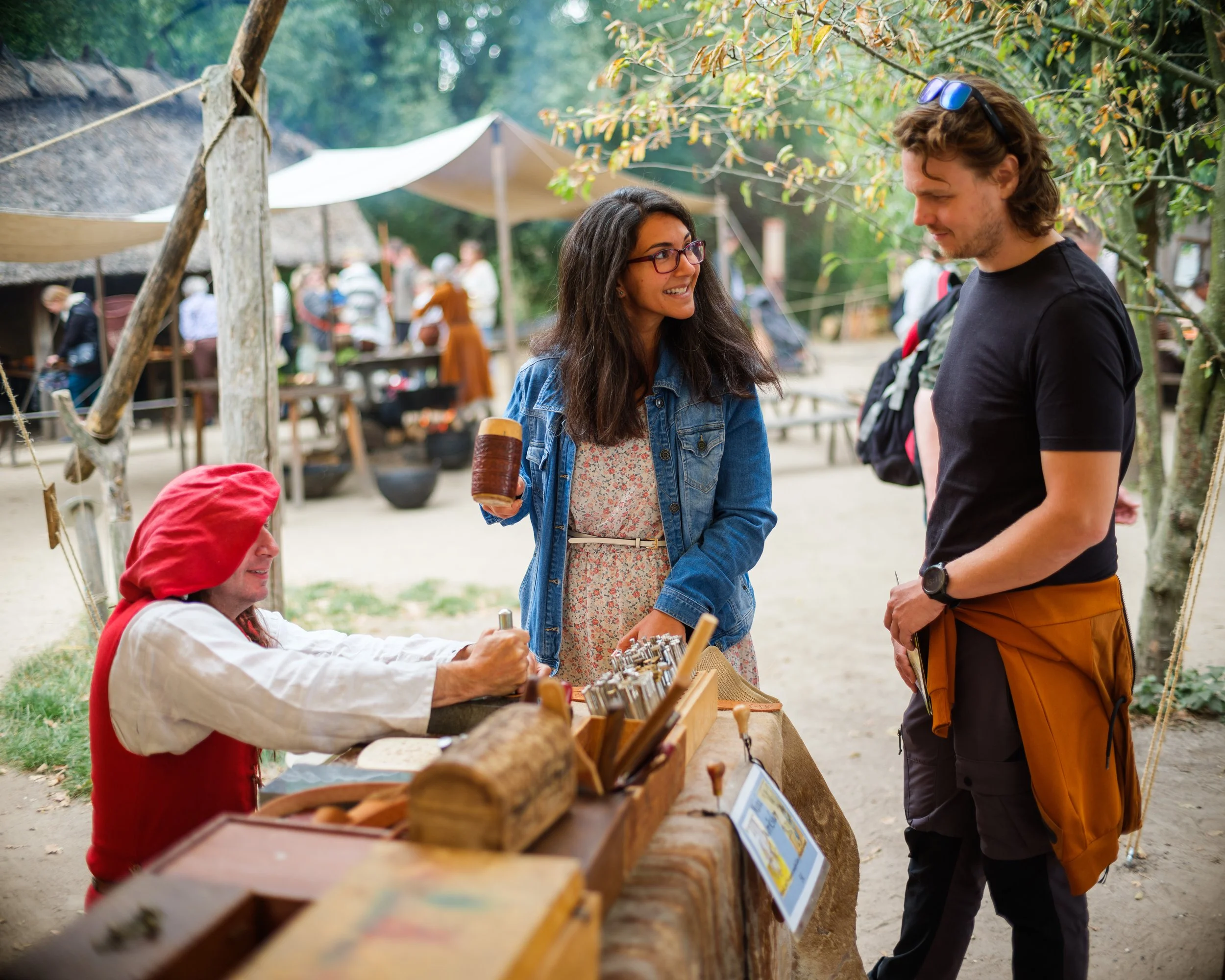 People browsing crafts at an outdoor market with trees and tents in the background.