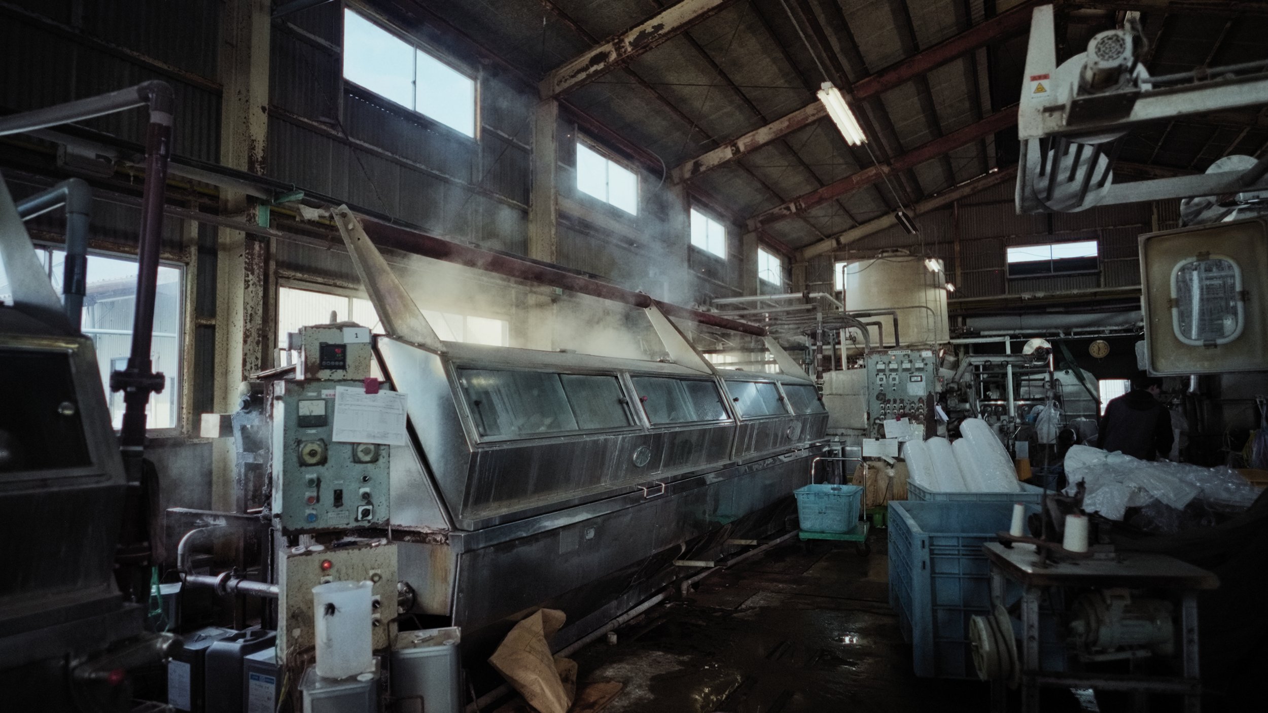 Industrial factory interior with machinery, pipes, and windows, some steam or smoke visible.