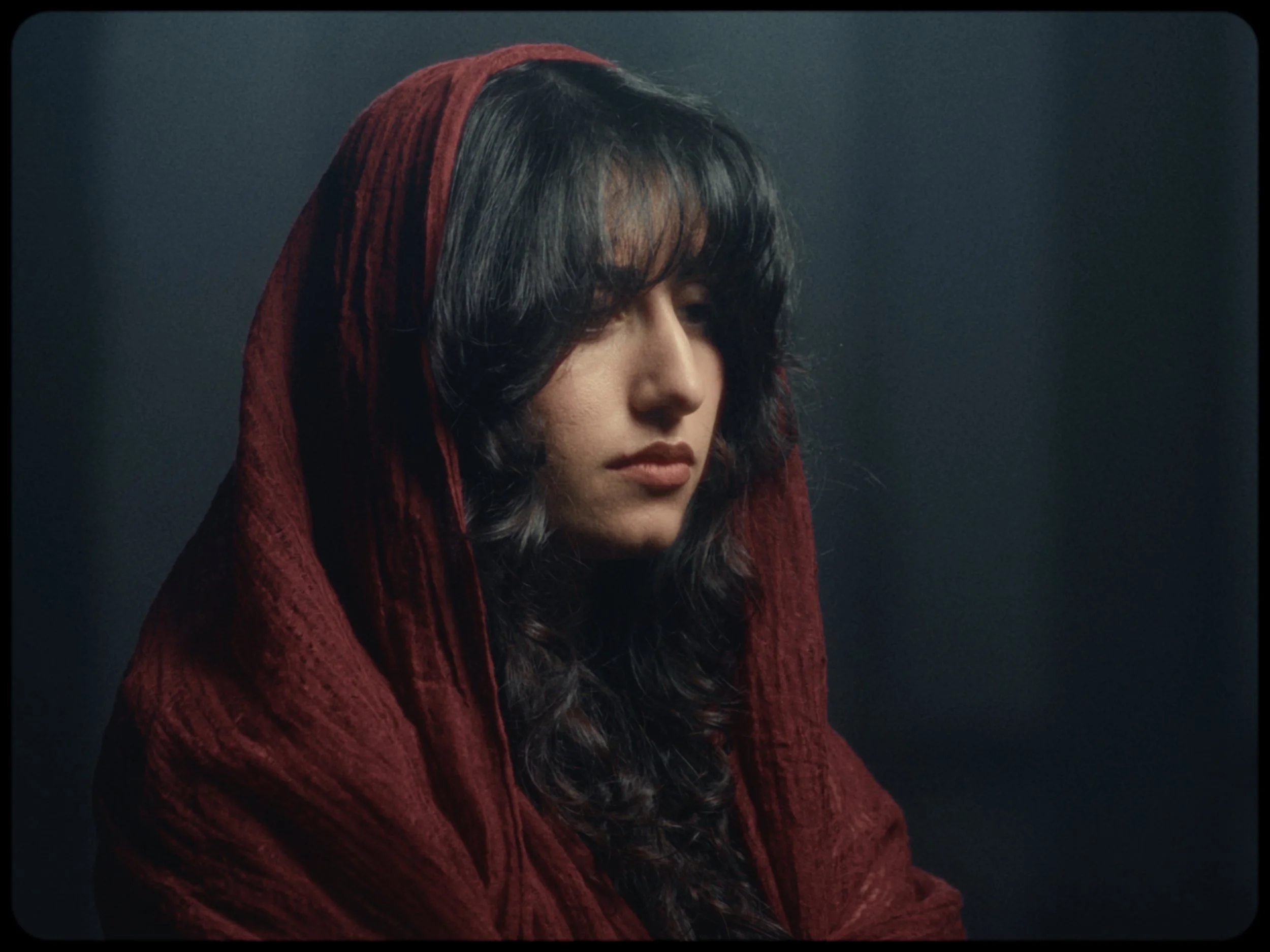 A woman with dark, wavy hair wearing a red hooded garment against a dark background, with eyes closed and a serene expression.