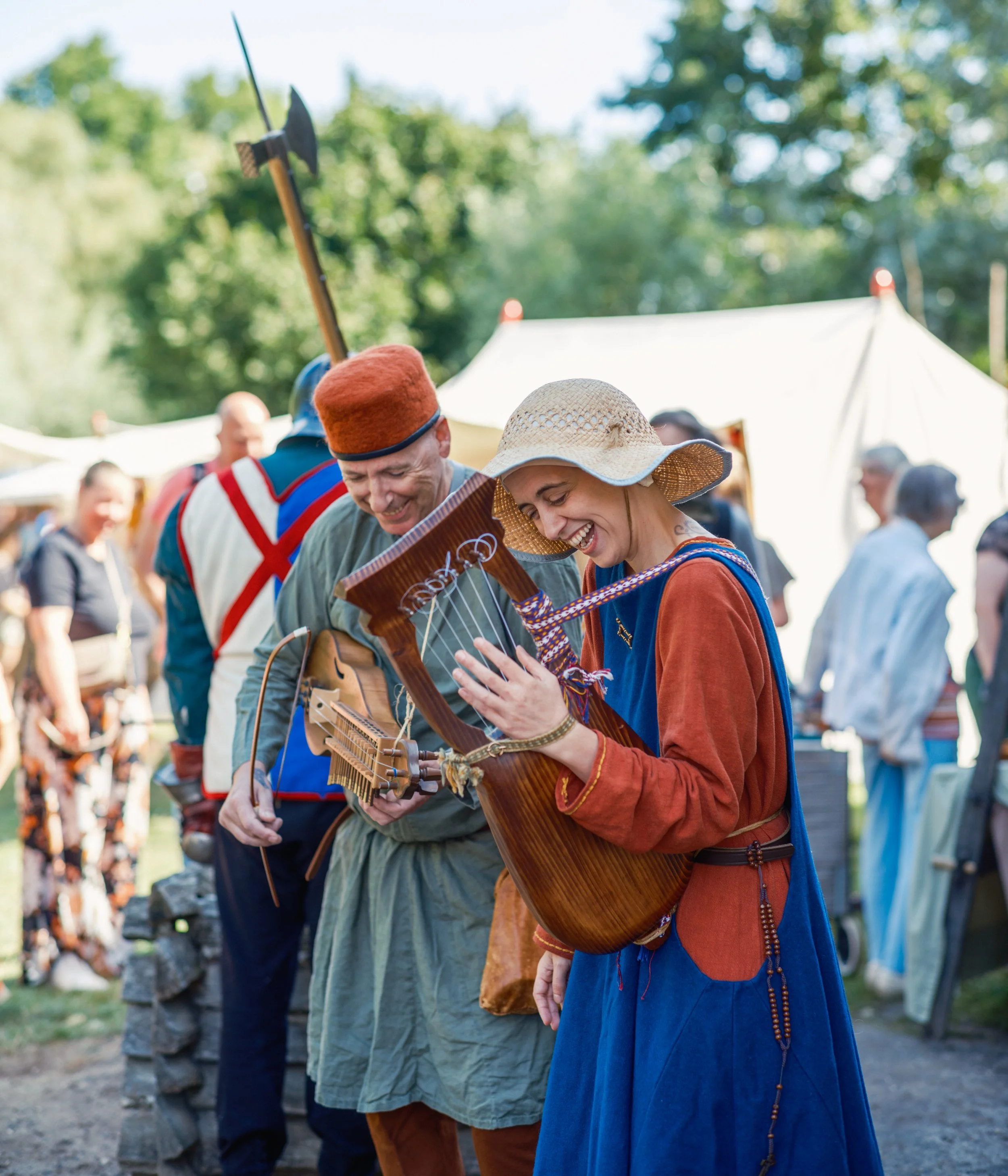 People in historical costumes, including a man with a gray tunic and a woman with a blue dress and wide-brimmed straw hat, engaging in a moment of music and laughter. Others are in the background, with a canopy tent and trees in an outdoor setting.