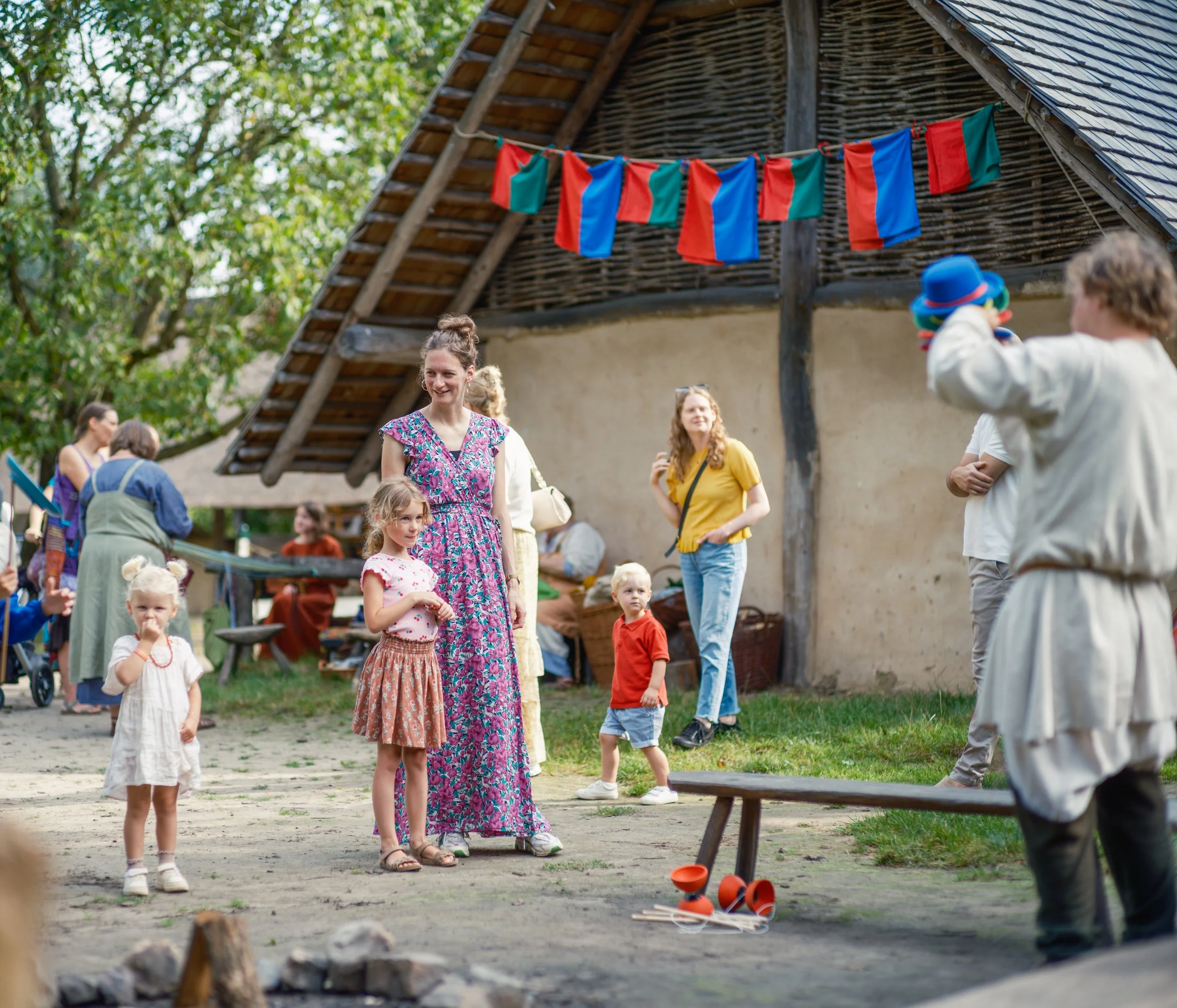 People standing and sitting outdoors near a wooden building with colorful flags hanging, children playing, and greenery in the background.