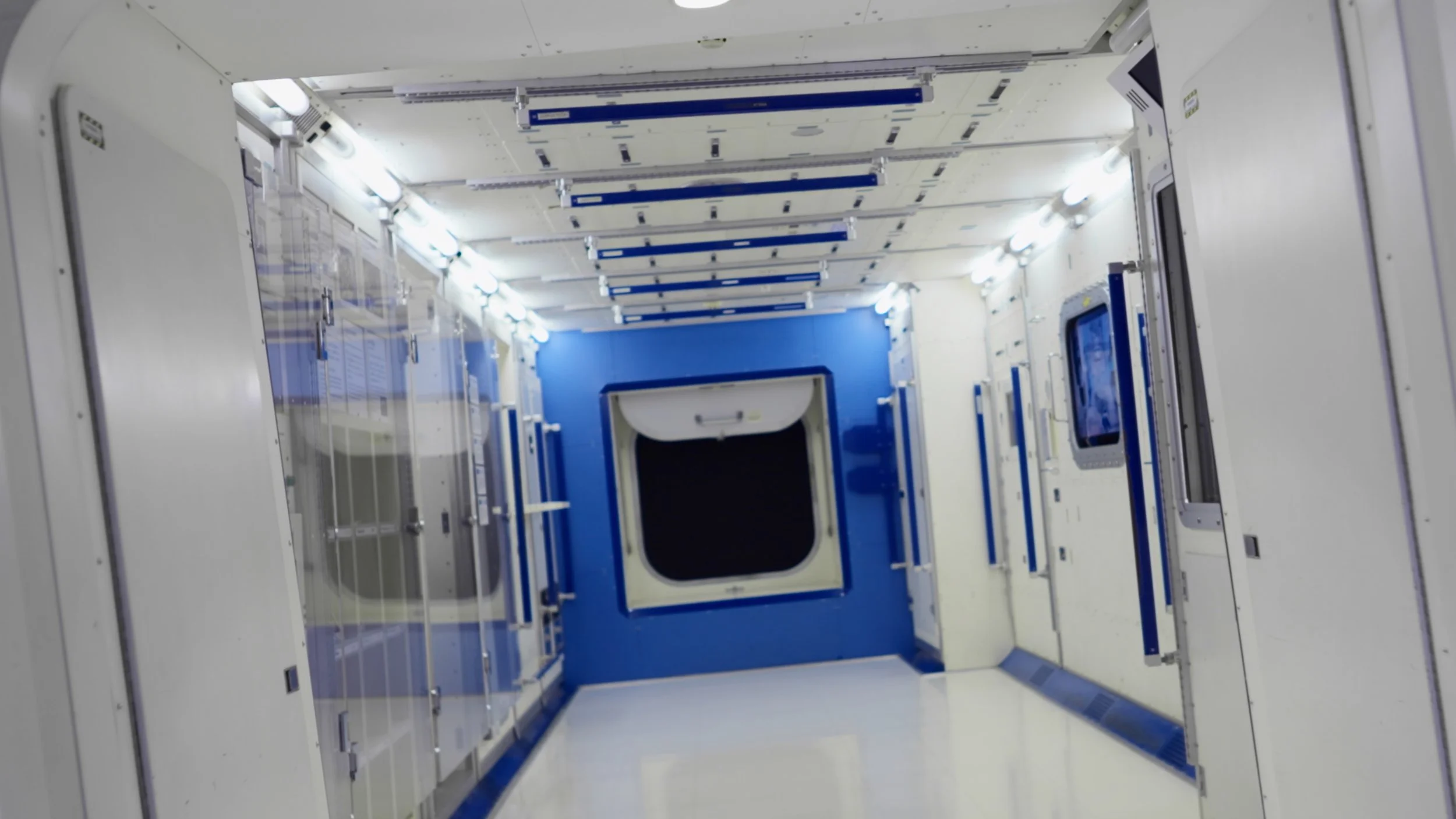 Interior view of a spacecraft or space station corridor with white panels, multiple lights, and a blue and white hatch at the end.