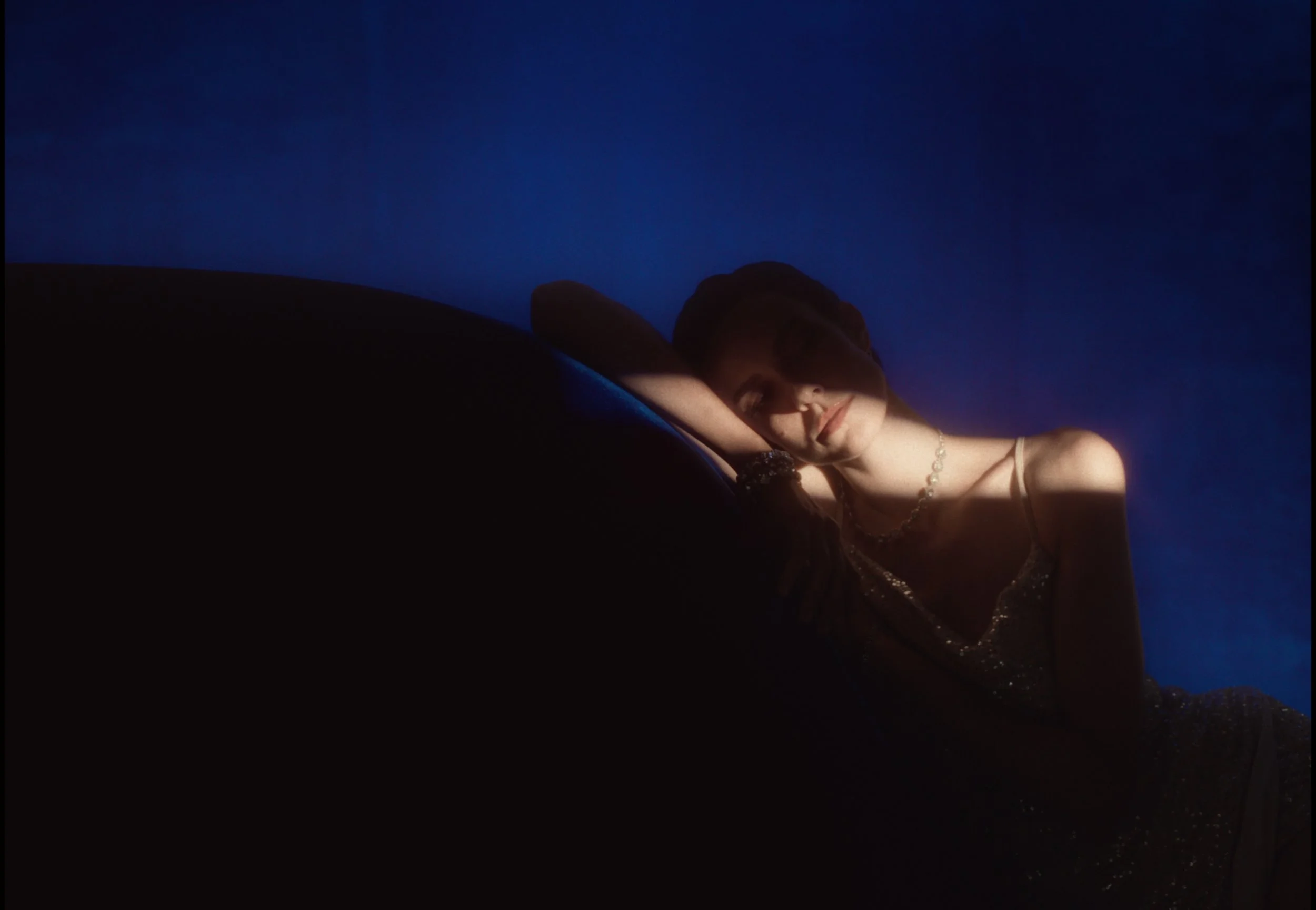 A woman with long hair and jewelry lying with her eyes closed in a dark, moody setting.