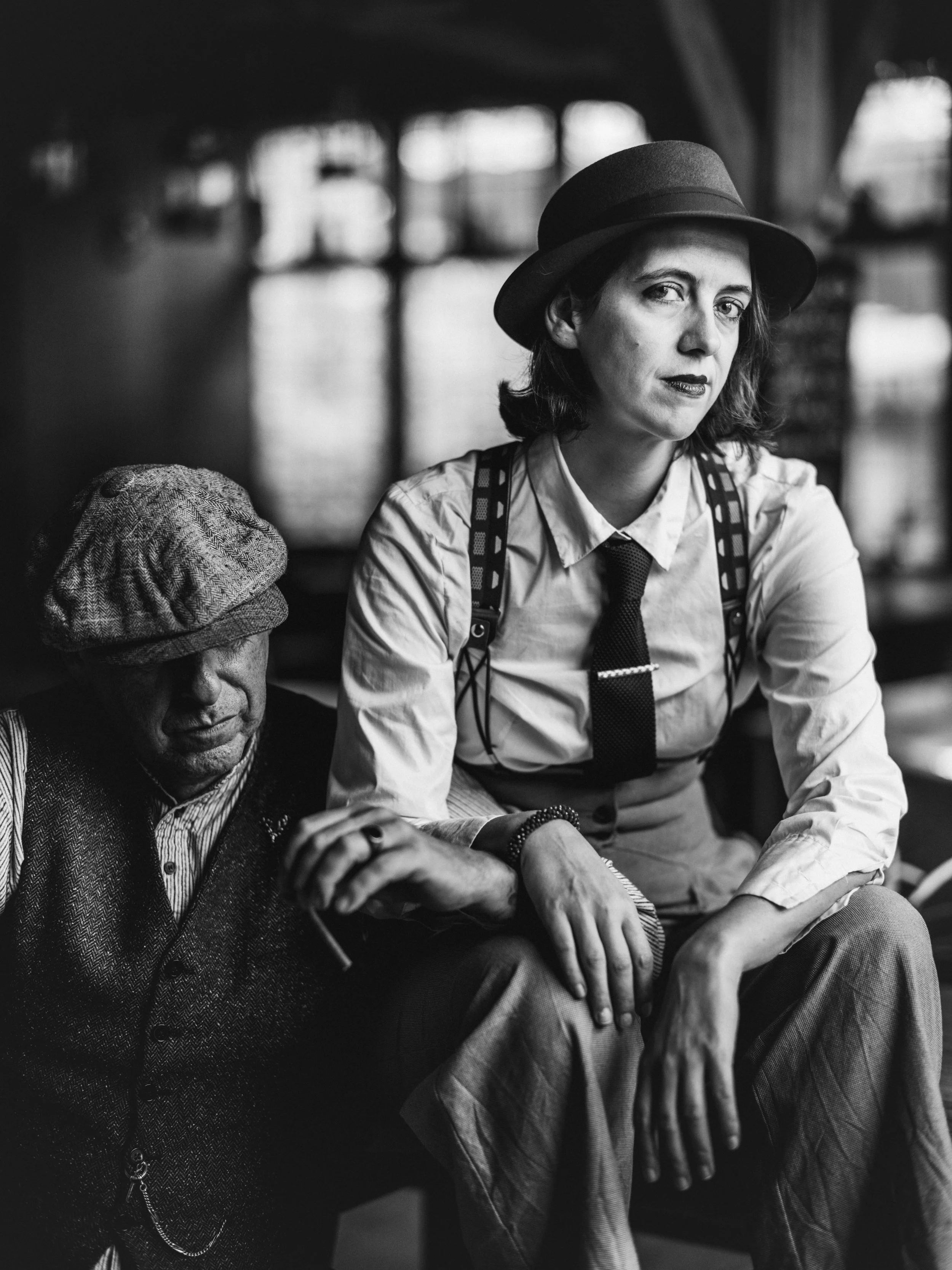 Black and white photo of a woman with a fedora hat sitting next to a man with a cap, holding hands, in a dimly lit environment with blurred background.