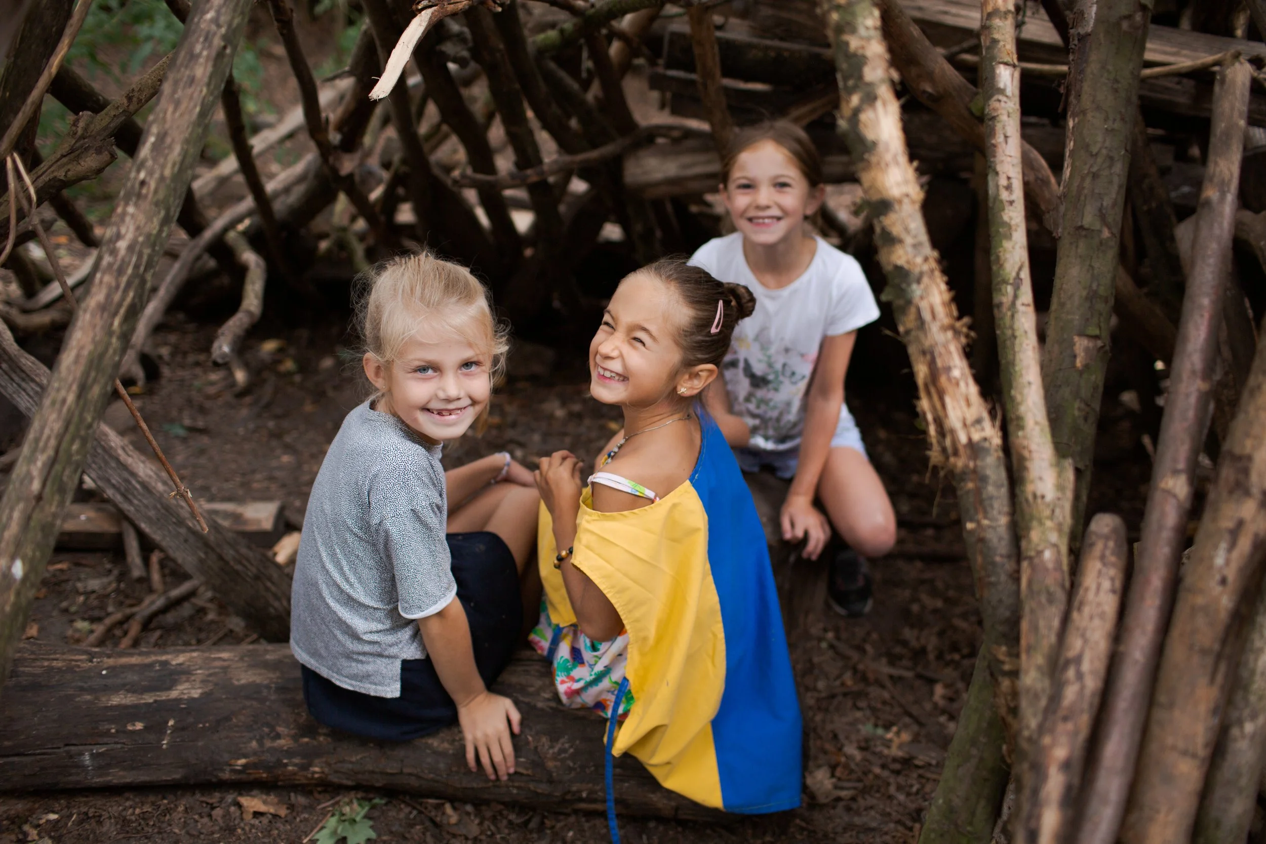 Four young girls smiling inside a small shelter made of branches in a wooded area.