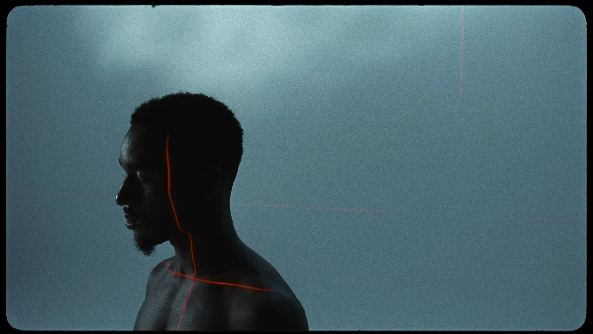 A side profile of a shirtless young man with short black hair, looking down, with glowing red lines tracing his face, neck, and shoulders, set against a dark, textured blue background.