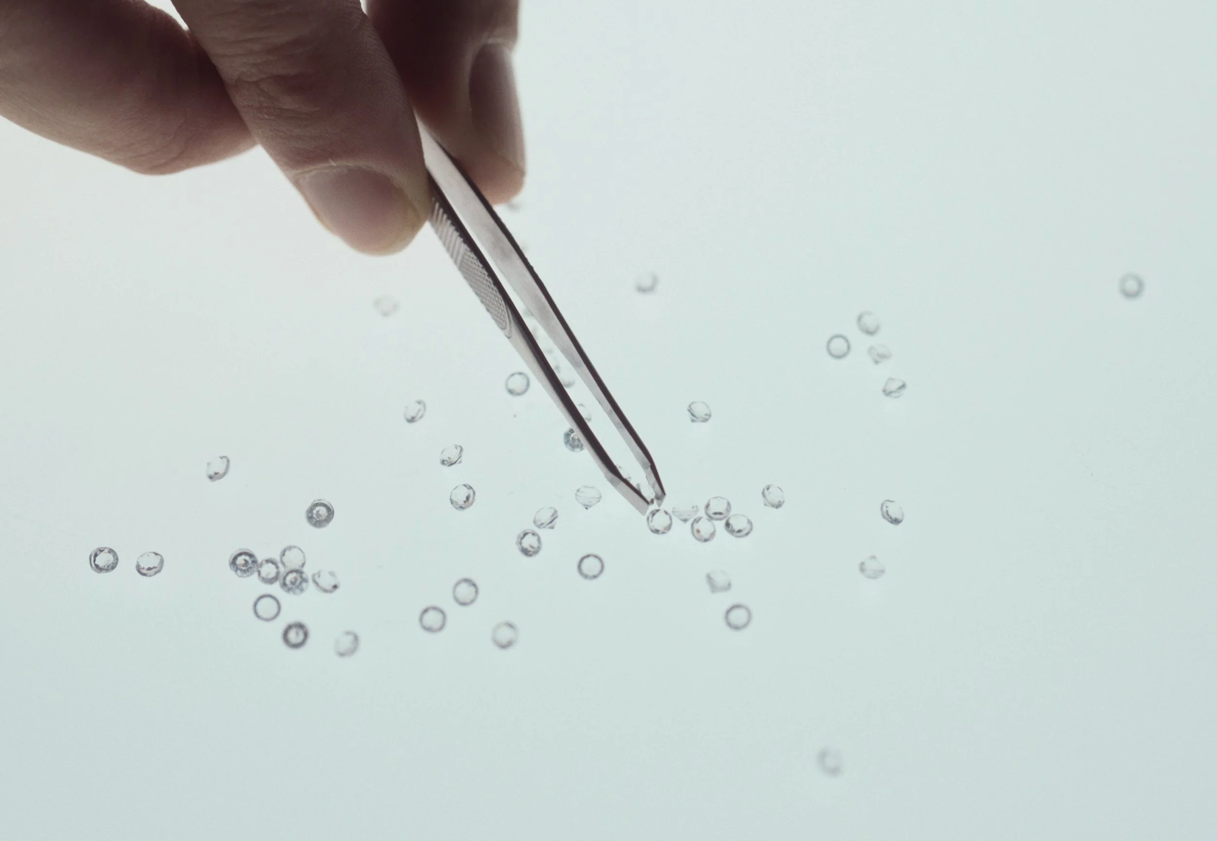 Close-up of a person's hand holding a pair of tweezers, picking up small clear glass beads on a plain surface.