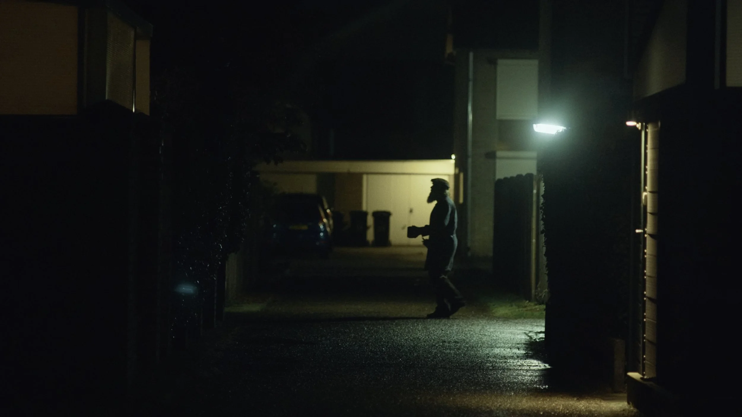 A person walking outdoors at night, holding a smartphone, with a house and trees in the background, illuminated by exterior lights.