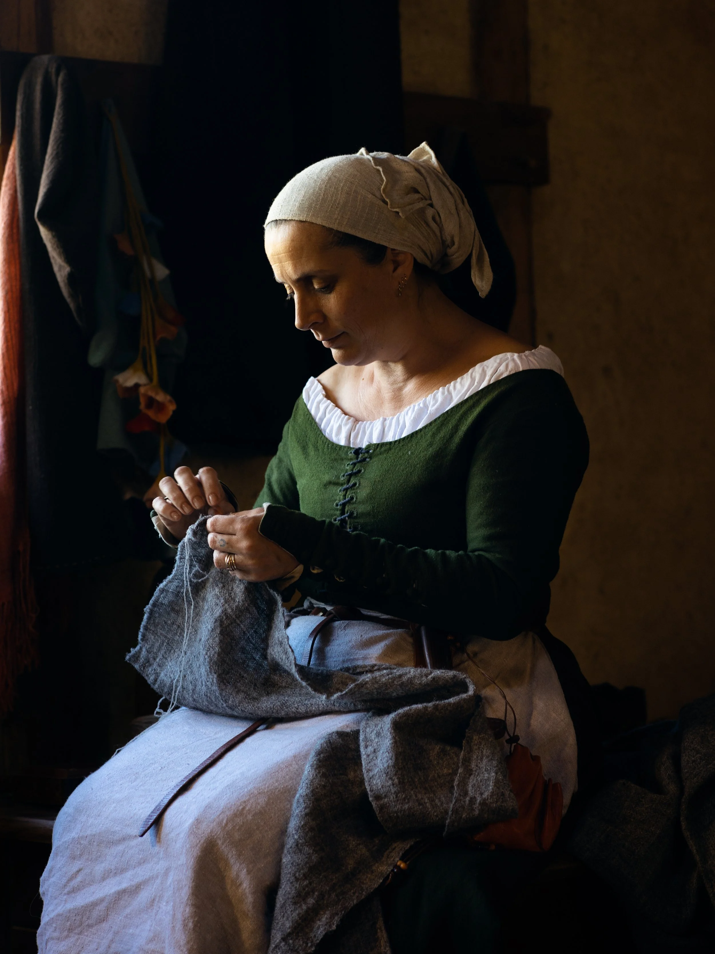 A woman dressed in historical clothing, including a gray headscarf and green dress, sitting in a dimly lit room, knitting with gray yarn.