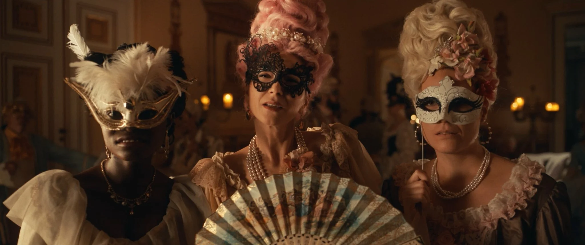 Three women in elaborate costumes wearing masks at a masquerade ball, with candles in the background.
