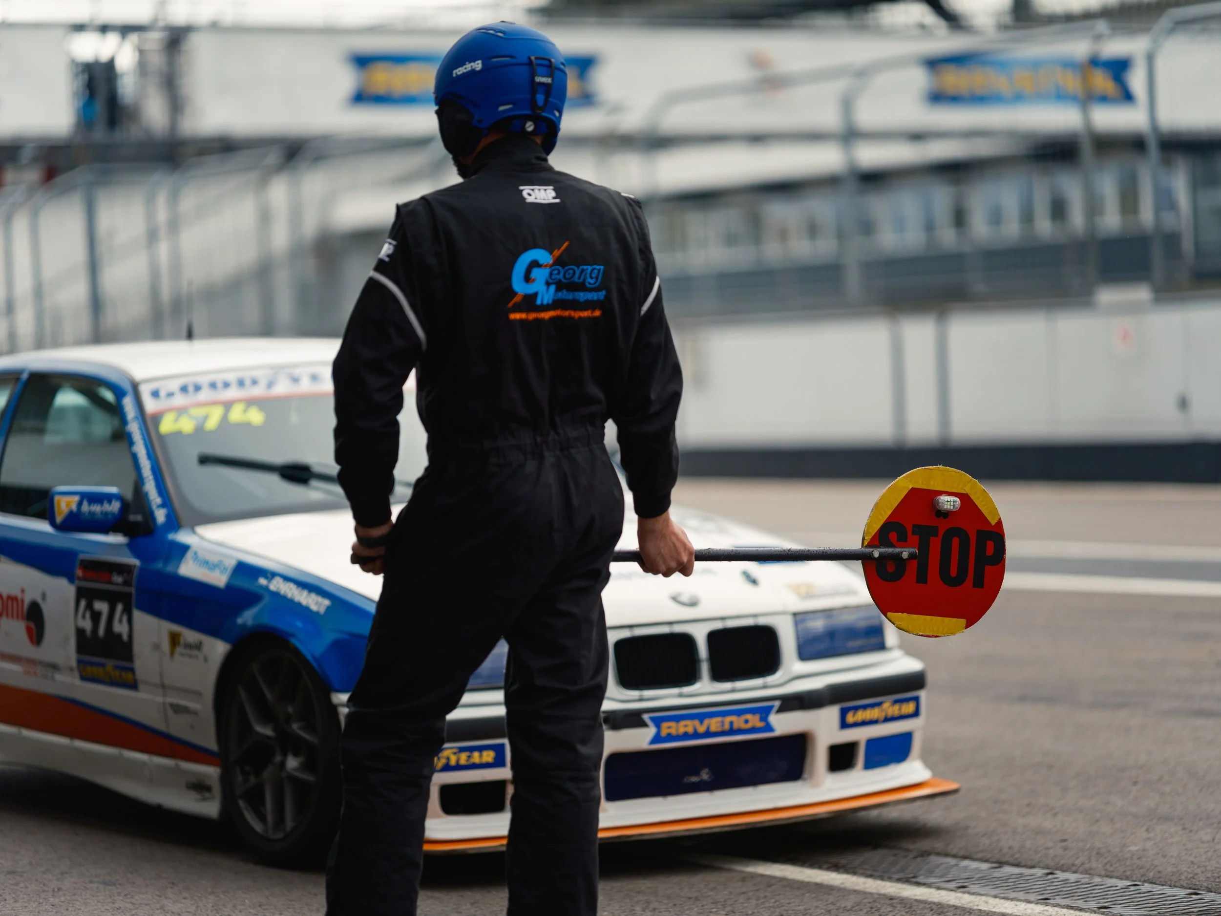 A race car driver wearing a blue helmet and black racing suit holding a stop sign standing near a blue and white race car at a racetrack.