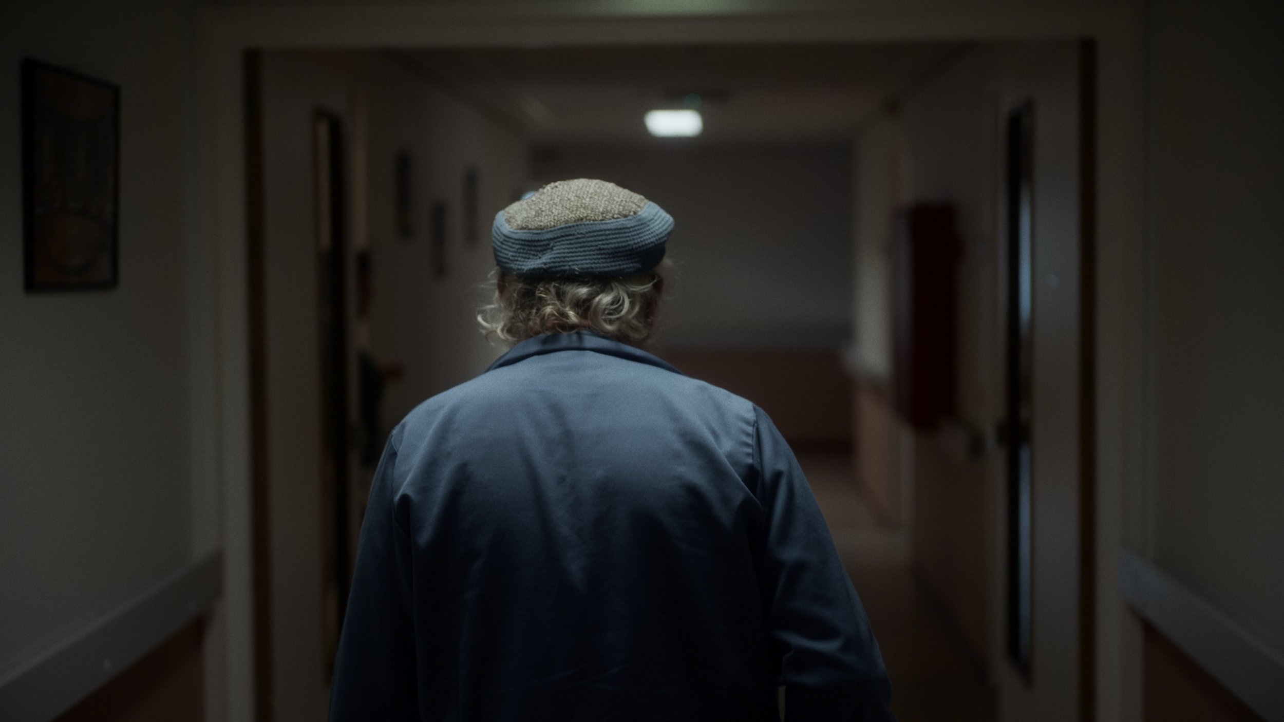 An elderly person with curly gray hair, wearing a blue cap and jacket, walks down a dimly lit hallway with framed pictures on the walls.