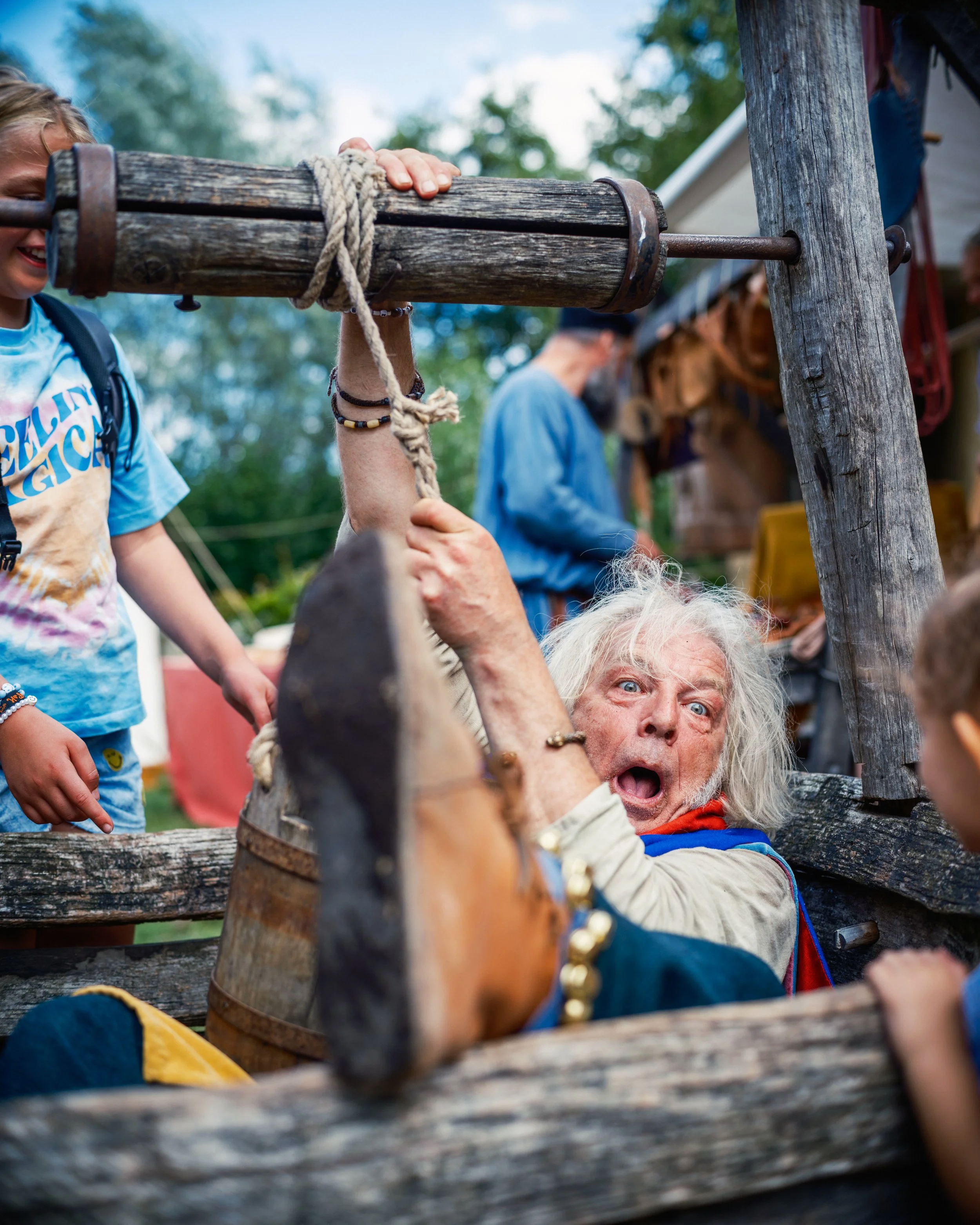 An elderly man with long white hair, wearing a colorful jacket, is looking up and appears surprised or excited while sitting behind a rustic wooden fence. A young girl with a backpack and a tie-dye shirt is smiling nearby. The setting looks outdoor w