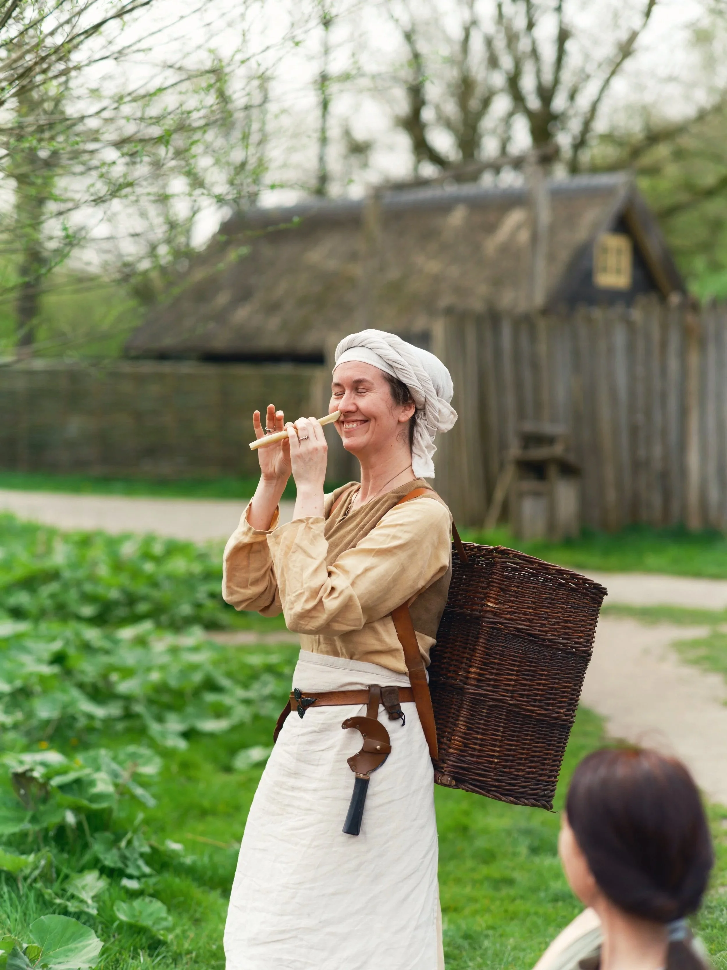 A woman dressed in medieval or fantasy-style clothing, smiling with eyes closed, holding a flute or pipe to her face, standing outdoors in a green area with trees and a wooden house in the background, with a person with dark hair in the foreground.