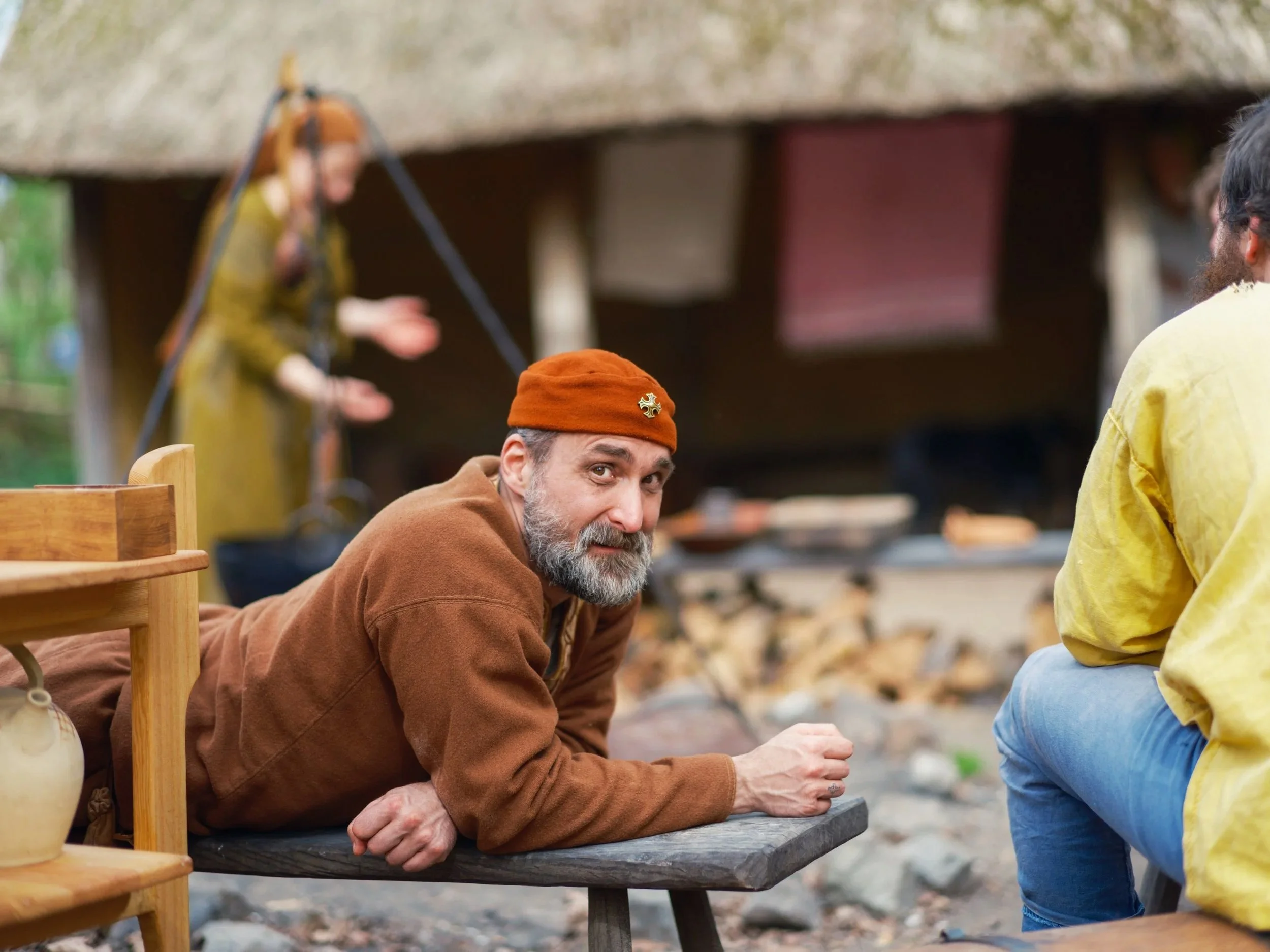 A man with gray hair and beard wearing a brown jacket and orange beanie with a cross pin, sitting at a wooden table outdoors, looking up and smiling at another person who is standing nearby wearing a yellow jacket, with a blurred background of a wood