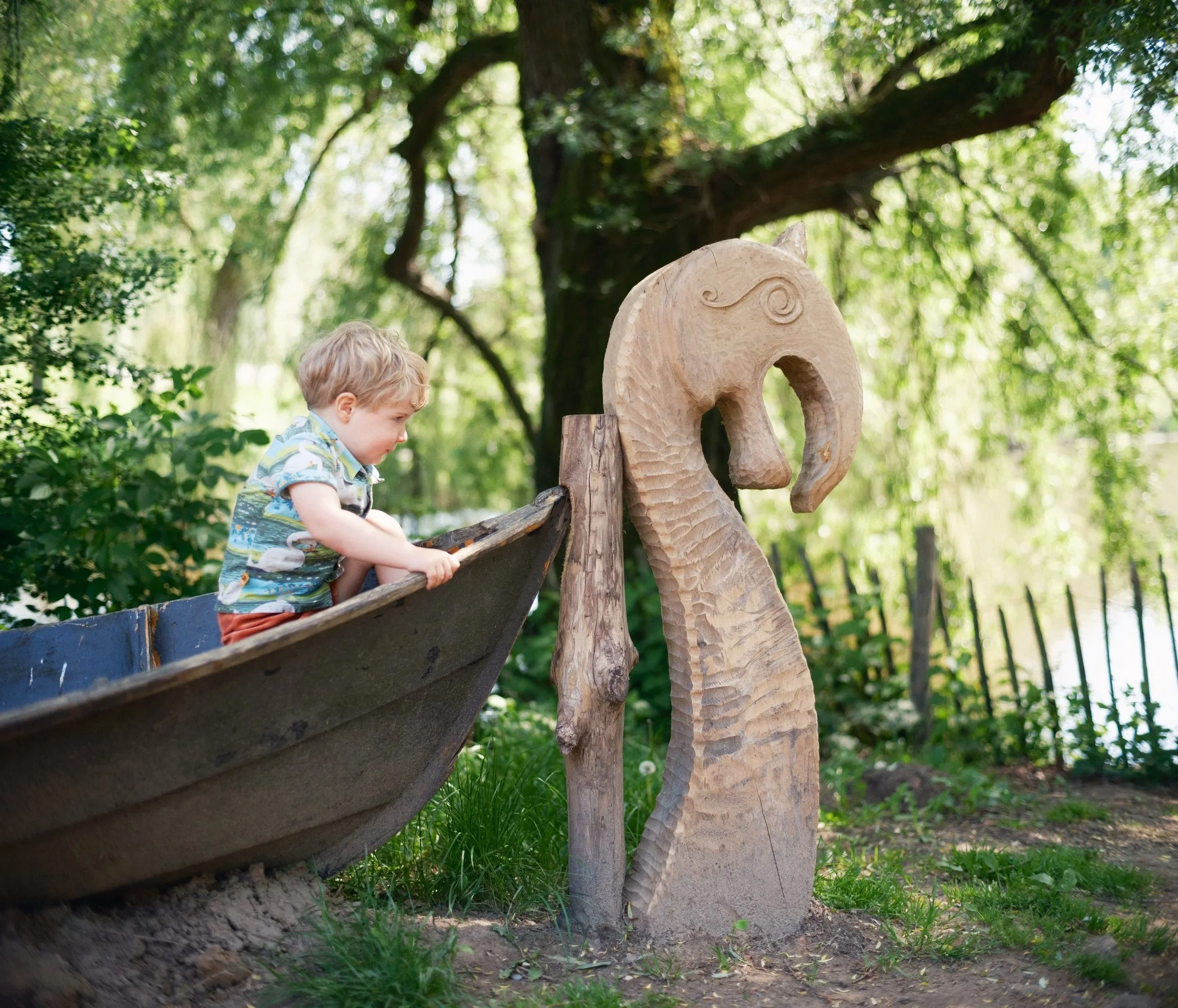 A young boy with curly blond hair leaning over a wooden railing outdoors, next to a carved wooden elephant statue in a lush green park or garden.