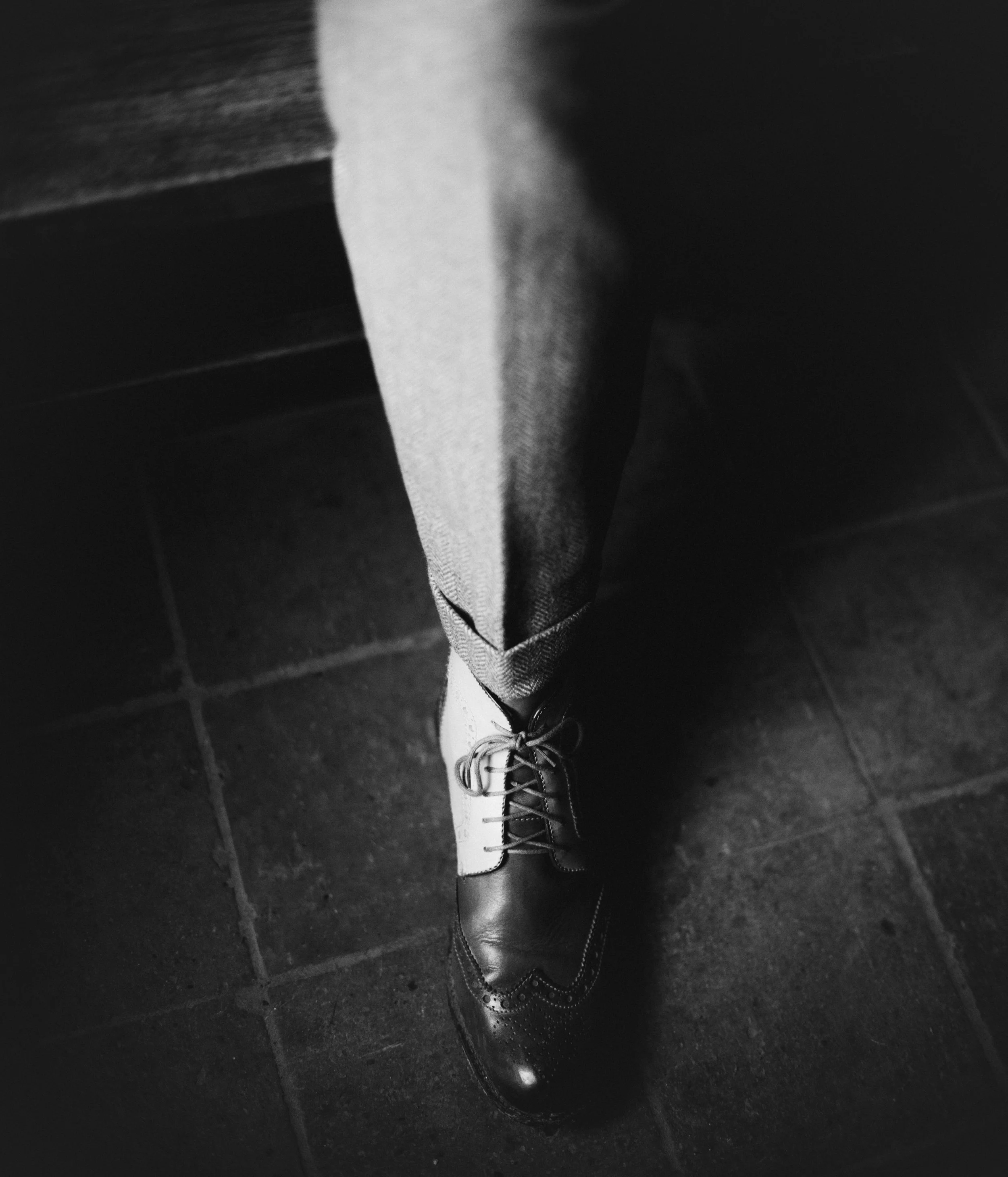 A person's legs wearing dress shoes and pants, standing on a dark tiled floor with a shadow cast on the side.