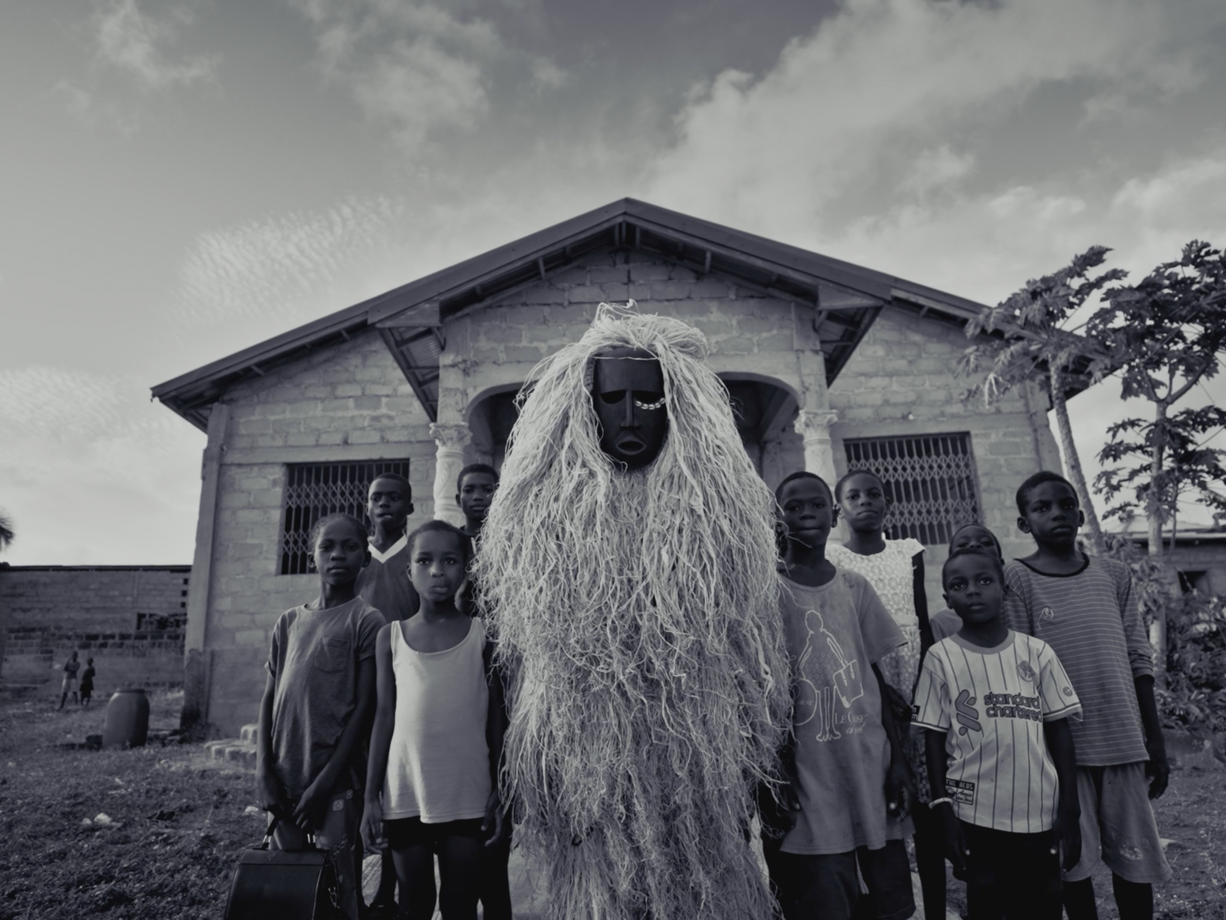 A person dressed in a traditional mask and costume with long hair stands in front of a group of children outside a house with a brick facade, trees, and a cloudy sky in the background.